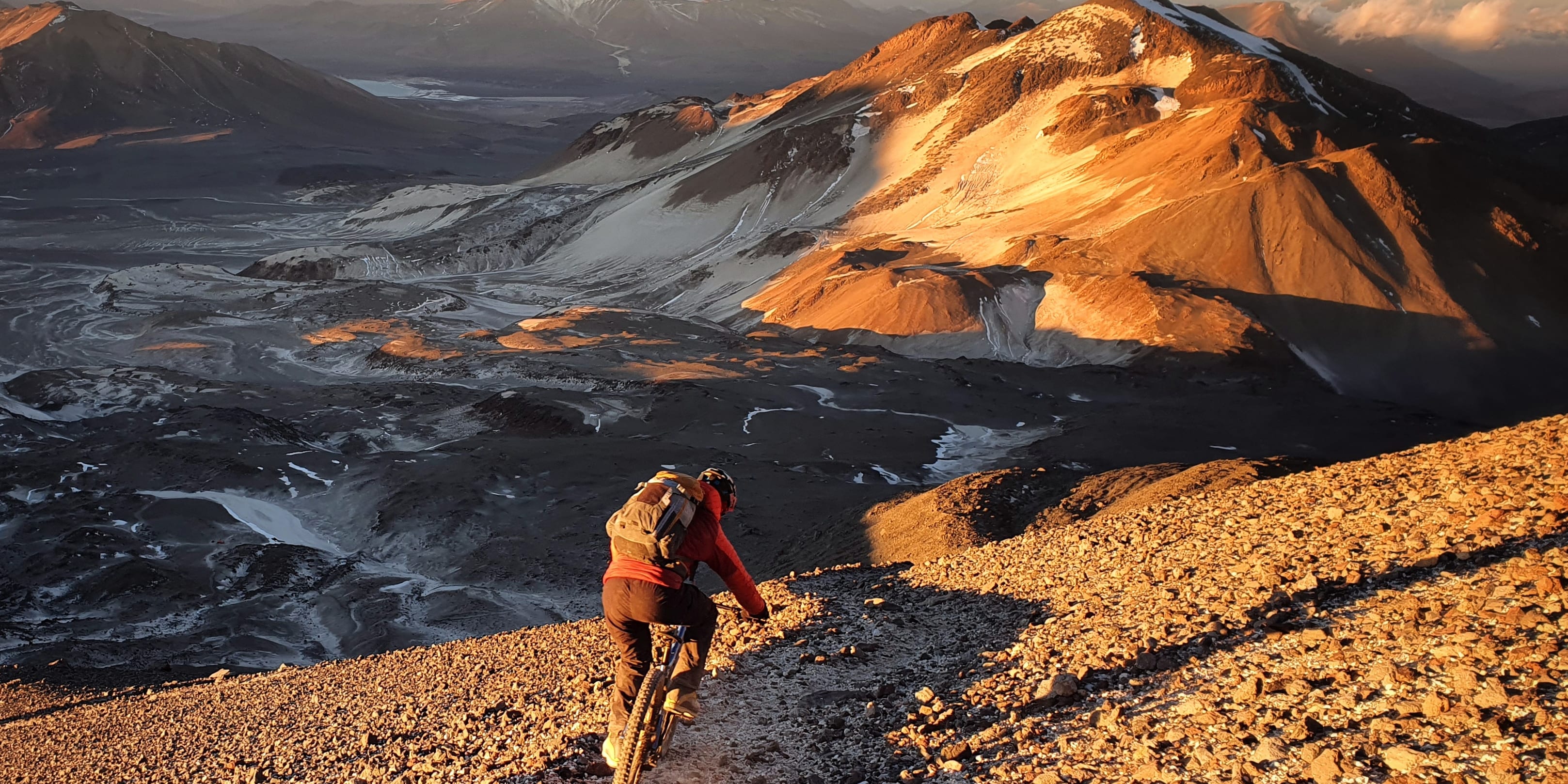 A person mountain biking down a rocky path in a mountainous landscape. The sun is setting behind the mountains, casting a warm glow on the scene. The biker is wearing a Norrøna jacket and riding a mountain bike. 
