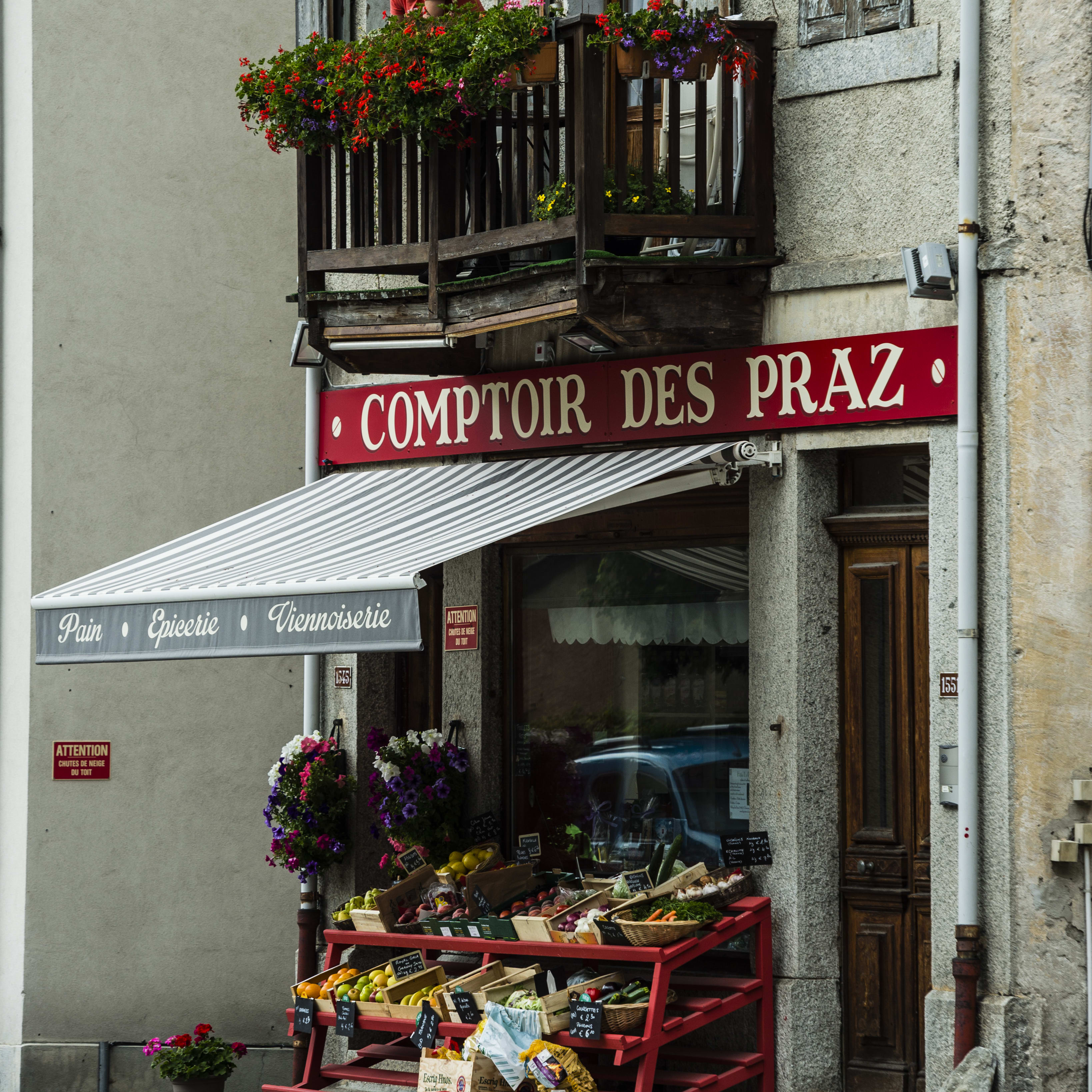 A man stands on a balcony overlooking a store called "Comptoir des Praz" with a striped awning. The store sells bread, groceries, and pastries. There is a red and white sign that says "Attention Chutes de Neige Du Toit" (Attention Snow Falling From The Roof) and a display of fresh produce in front of the store. 
