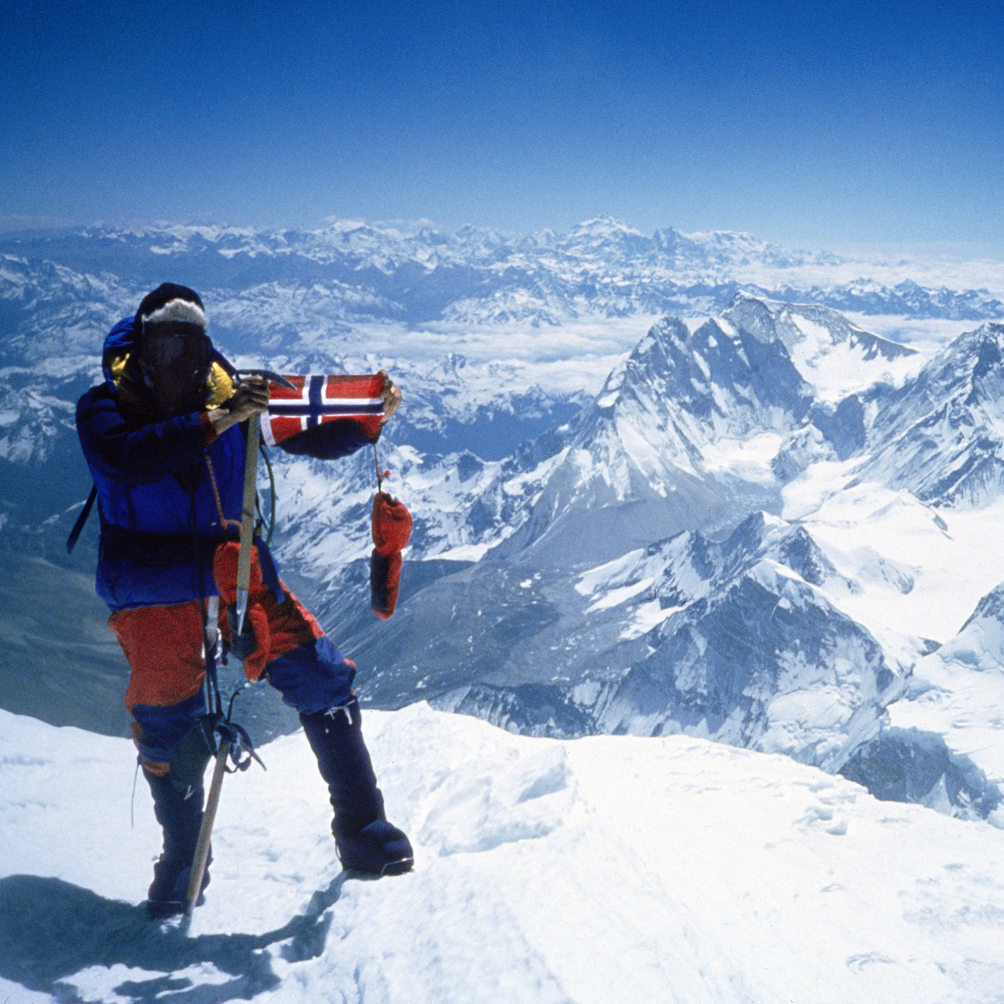 A climber in Norrøna gear stands on a snowy mountain peak, holding a Norwegian flag. Mountains and a clear blue sky are visible in the background.
