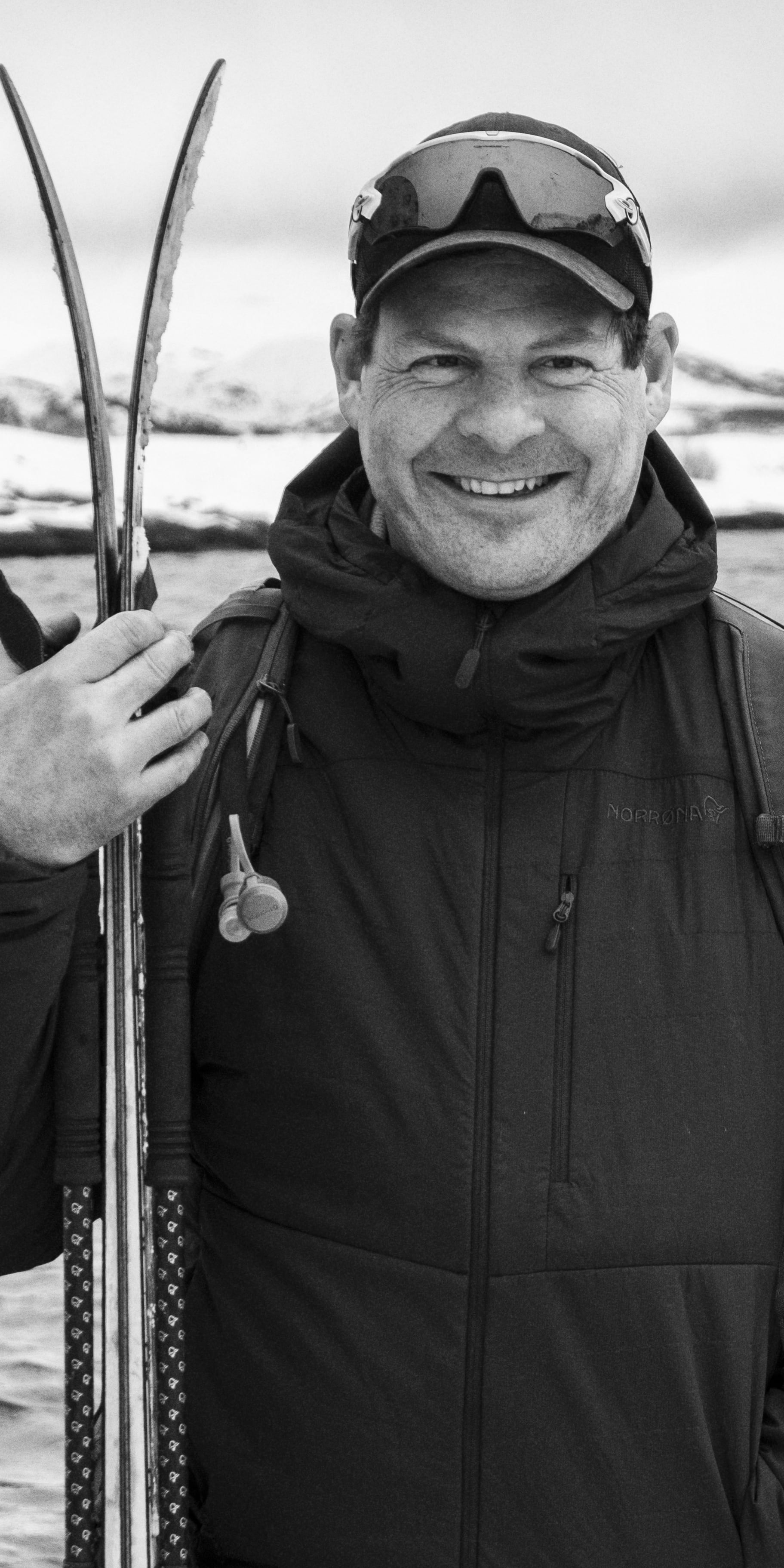 A man wearing a black Norrøna jacket and a baseball cap holds two skis in his hand, standing in front of a body of water. 
