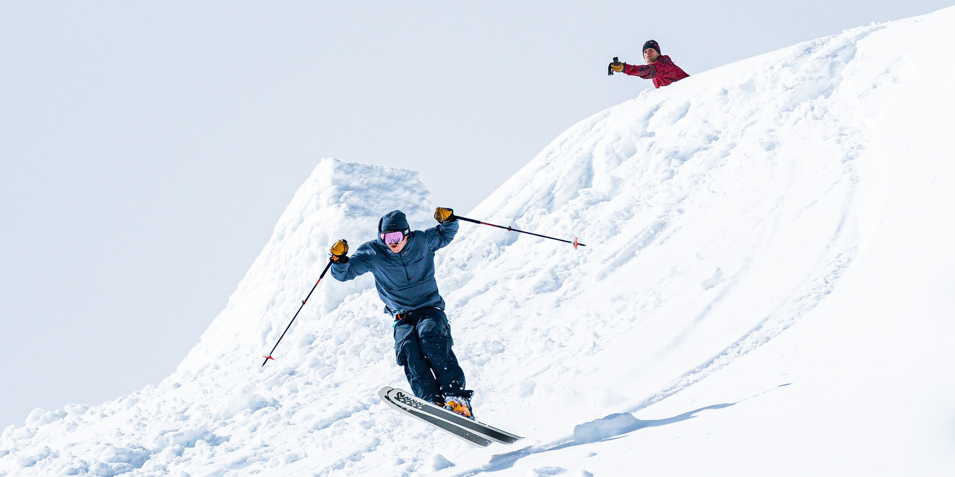 A skier wearing Norrøna clothing jumps off a snowdrift.