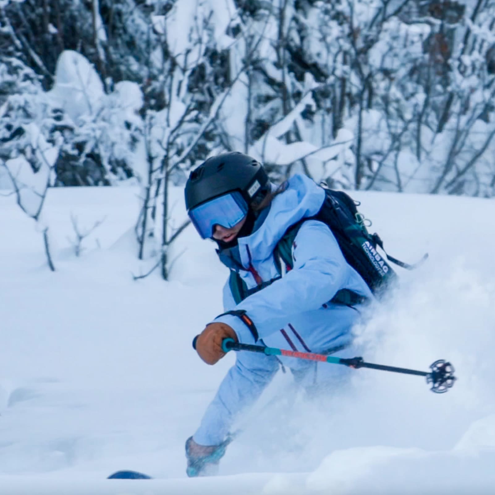 A skier wearing a Norrøna jacket, helmet, and goggles, carves through deep powder snow in a snowy forest.