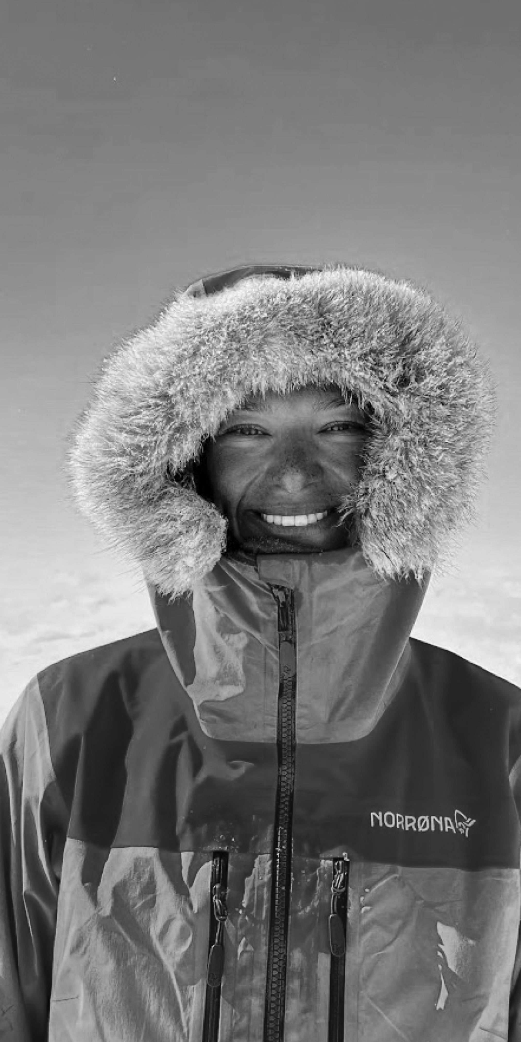A smiling man wearing a black and white Norrøna jacket with a fur hood, standing outside. 
