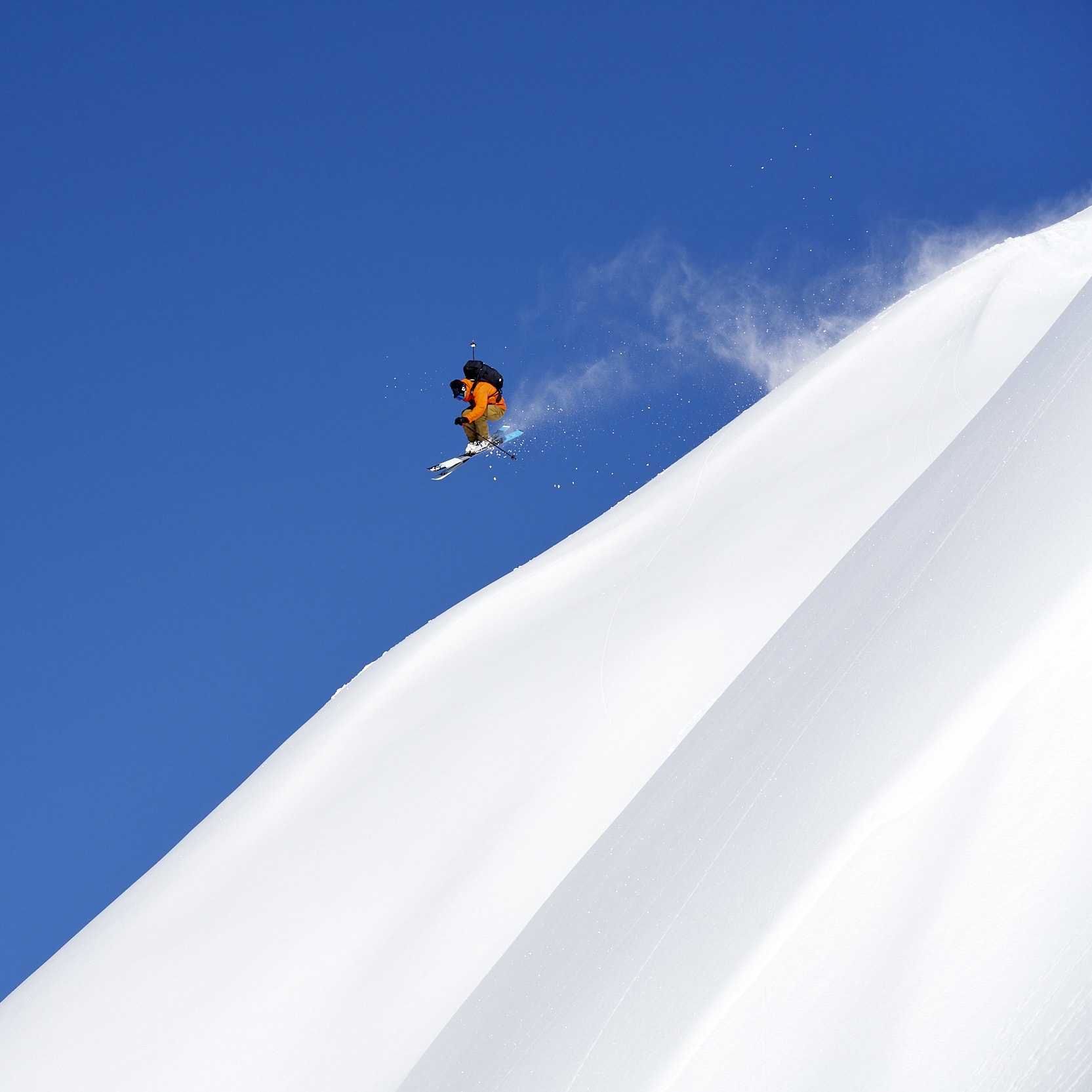 A skier in orange Norrøna outerwear jumps off a snow-covered mountain against a clear blue sky.