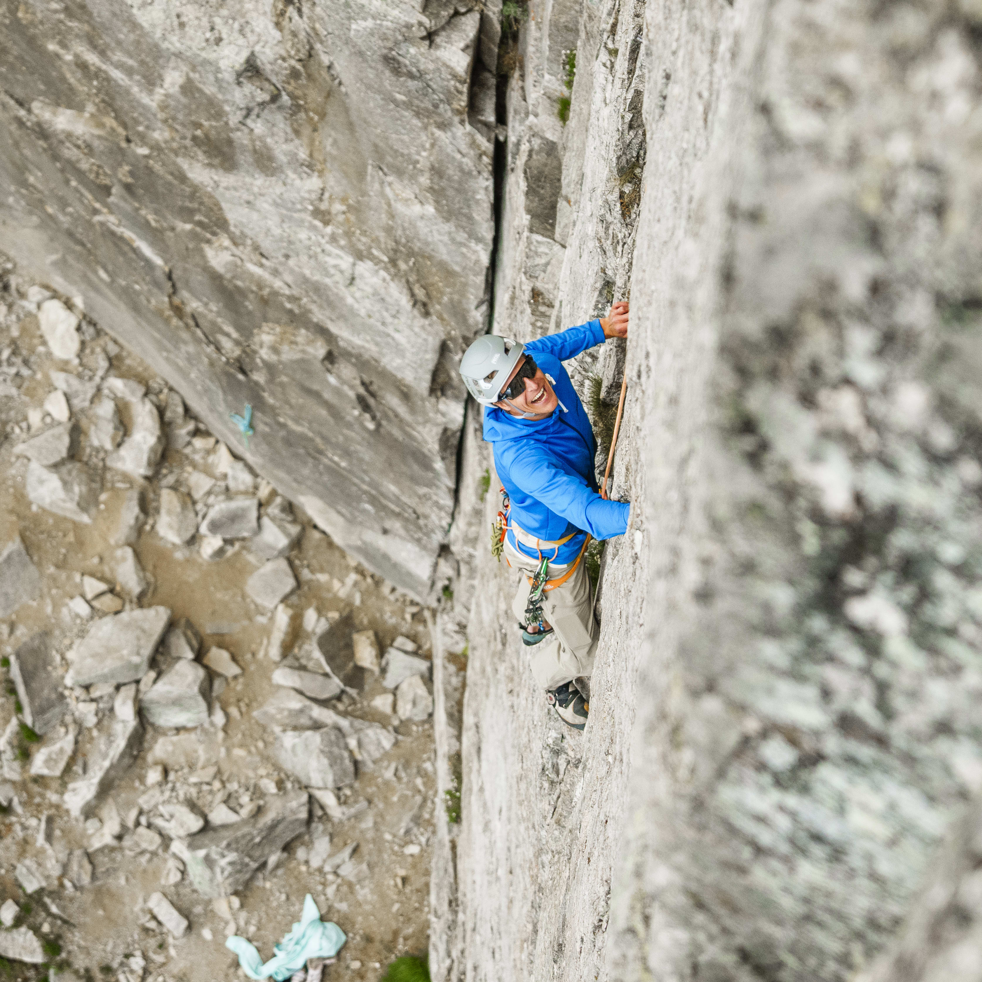 A rock climber smiles in a Norrøna blue jacket, helmet, and harness, ascending a steep, textured rock face with safety ropes and gear.