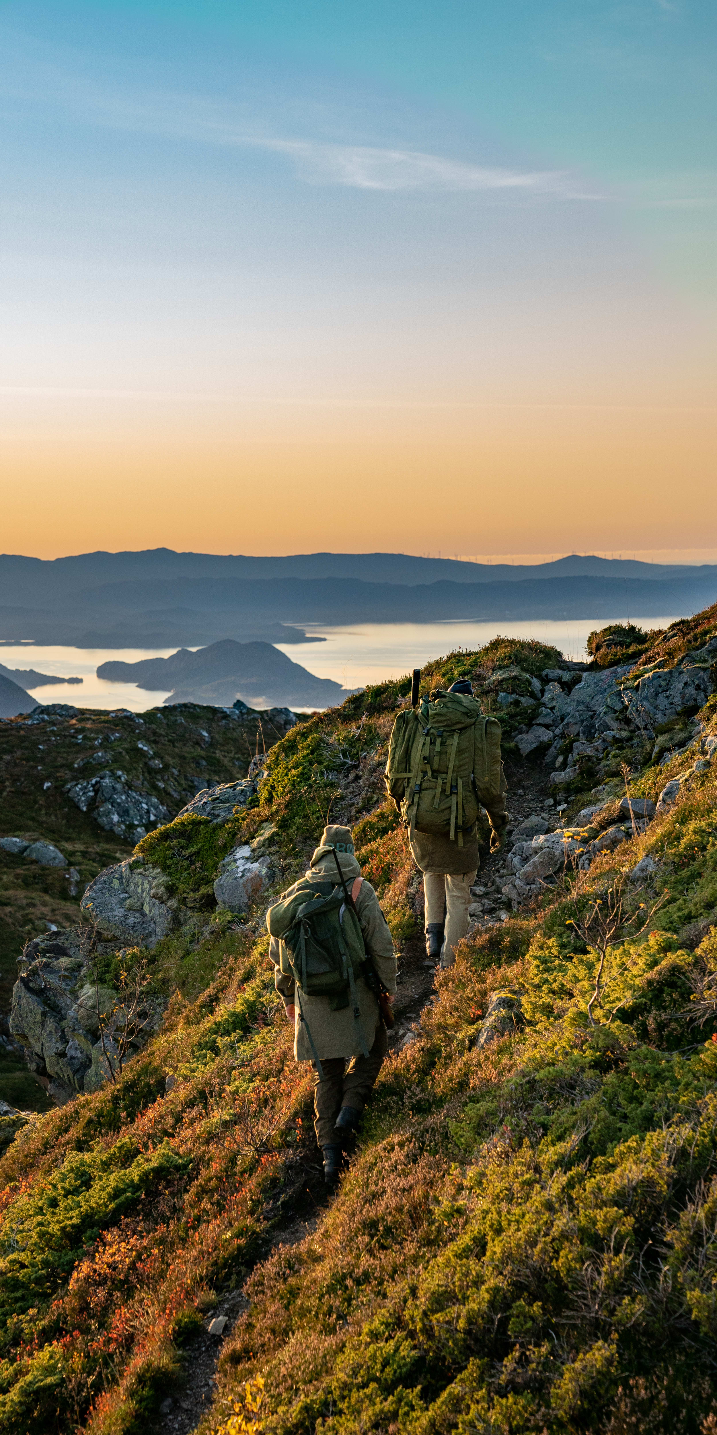 Two hikers wearing Norrøna backpacks walk on a mountain path overlooking a fjord at sunset. 
