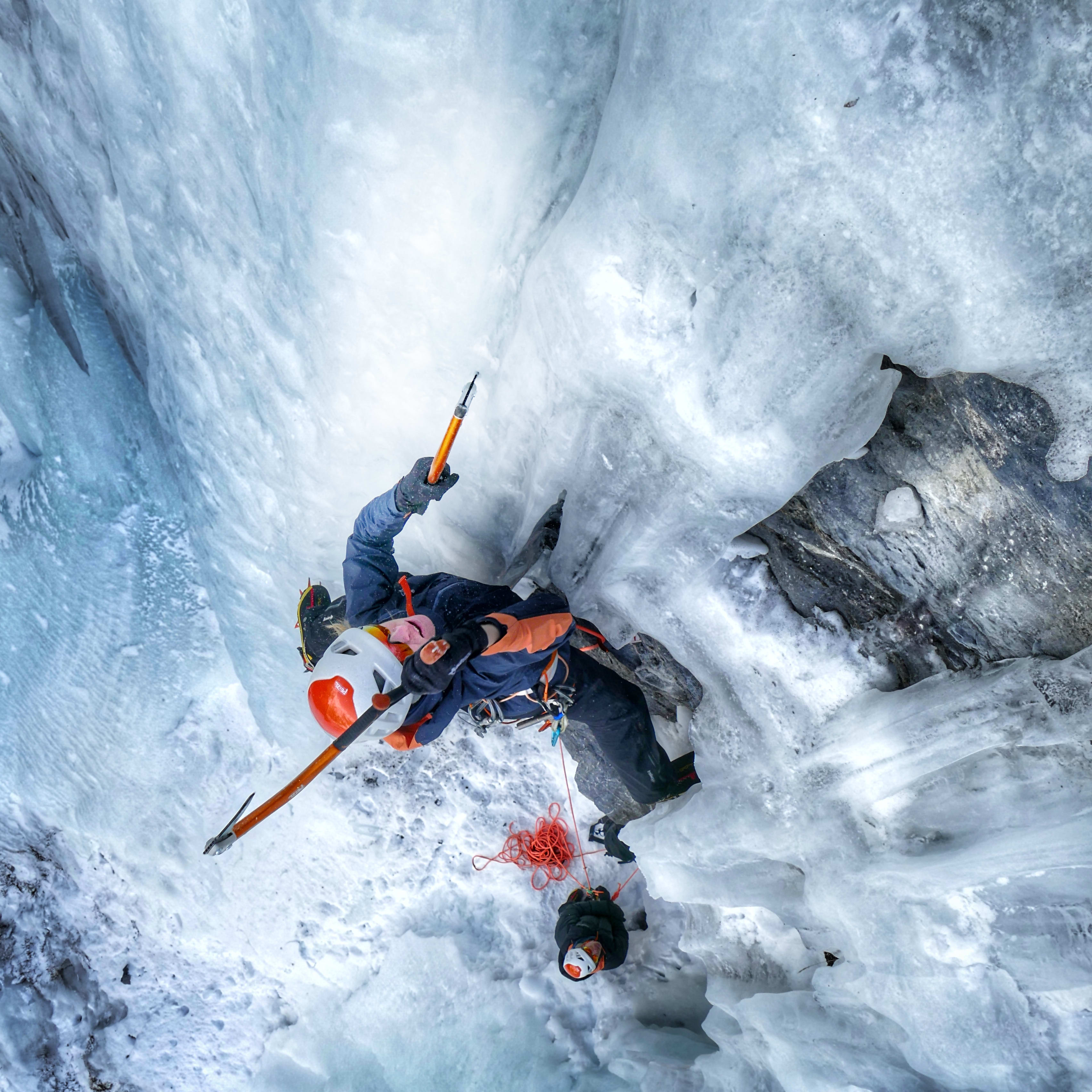 Eye-level view of ice climber using ice axes on a frozen waterfall, wearing Norrøna jacket and helmet.
