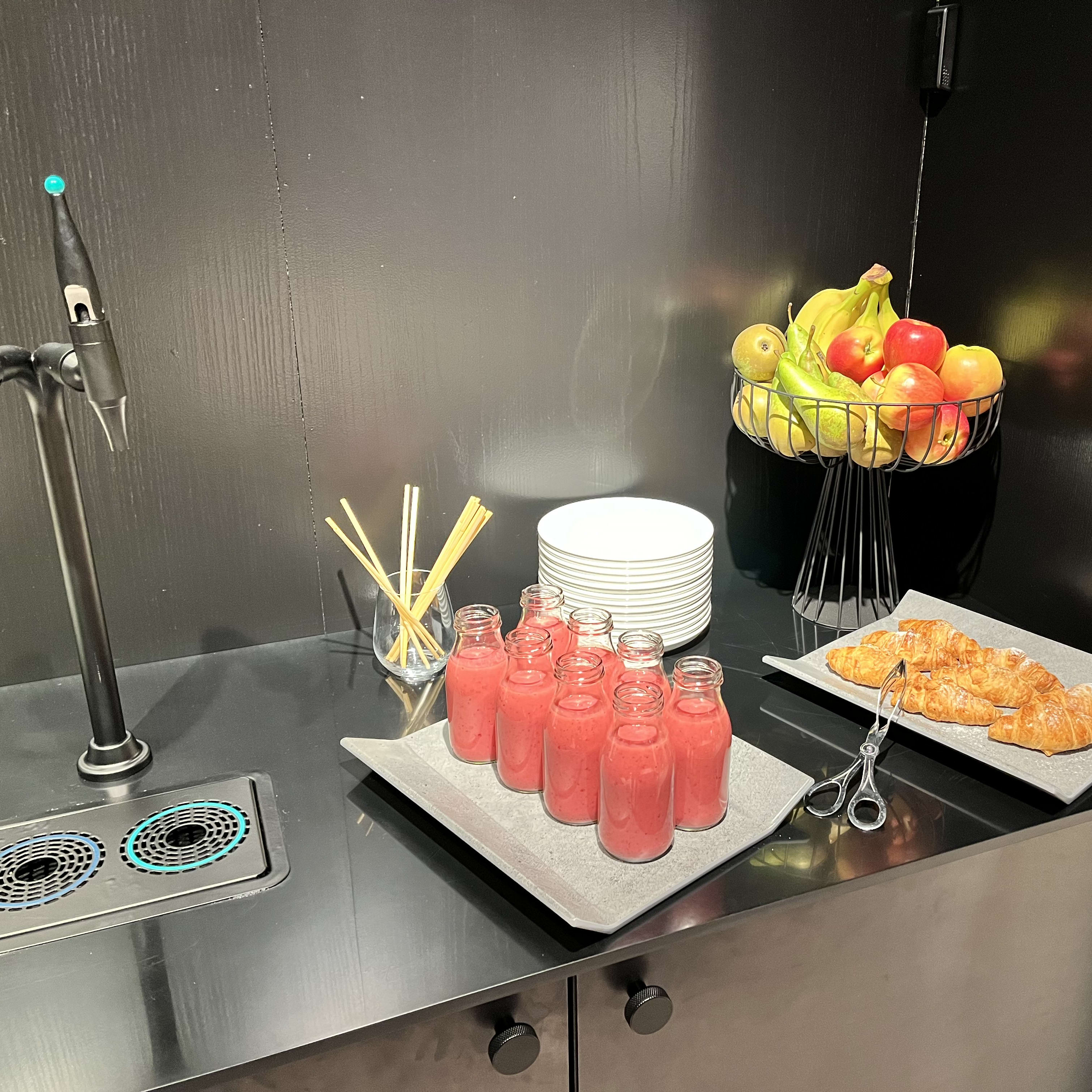 A black counter with a water tap, six glass bottles of pink smoothie, a stack of white plates, a bowl of fruit, and a plate with croissants and a pair of silver scissors. All clothing present in the image is from Norrøna.