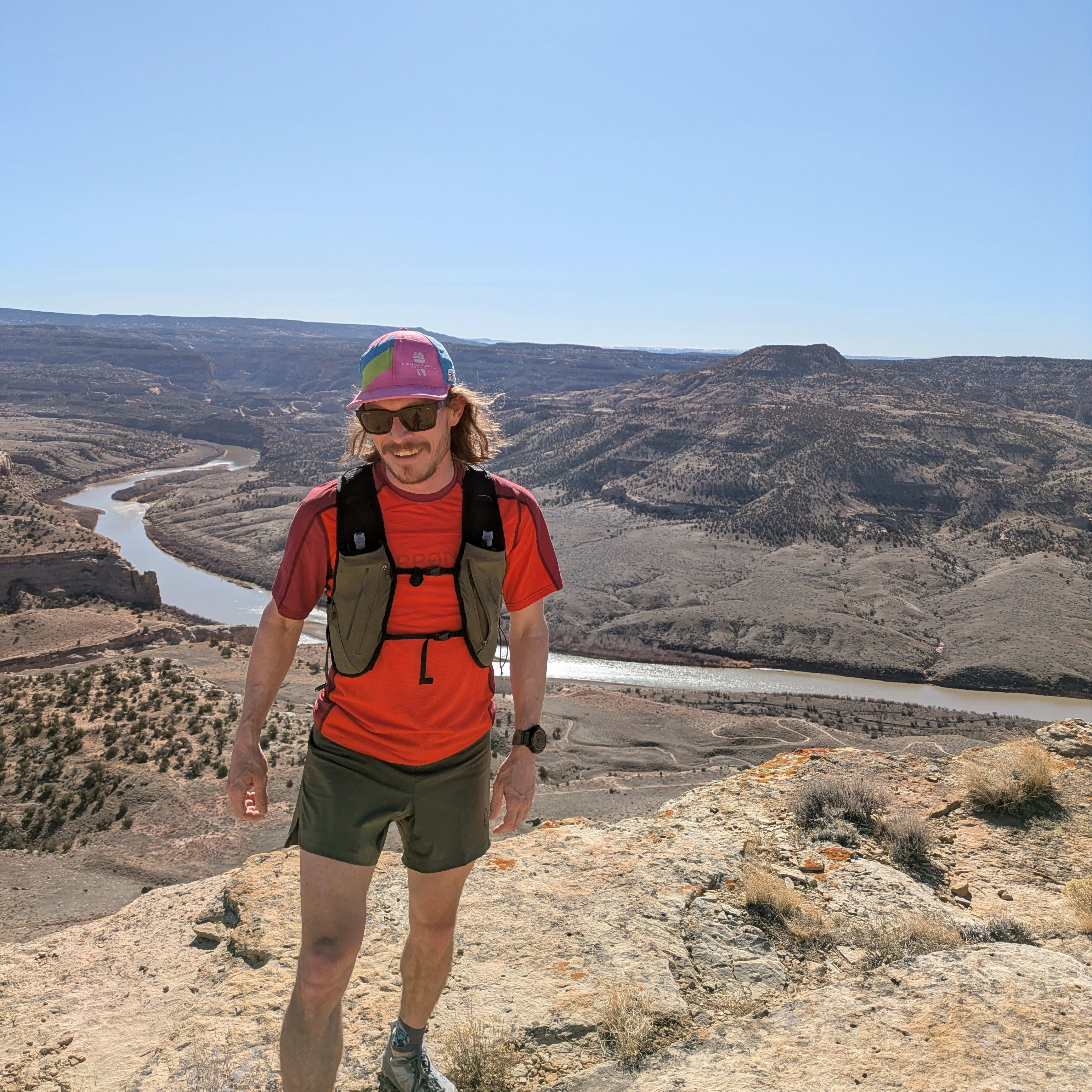 A man wearing Norrøna shorts and a shirt stands on a rocky cliff overlooking a river in a desert landscape. 
