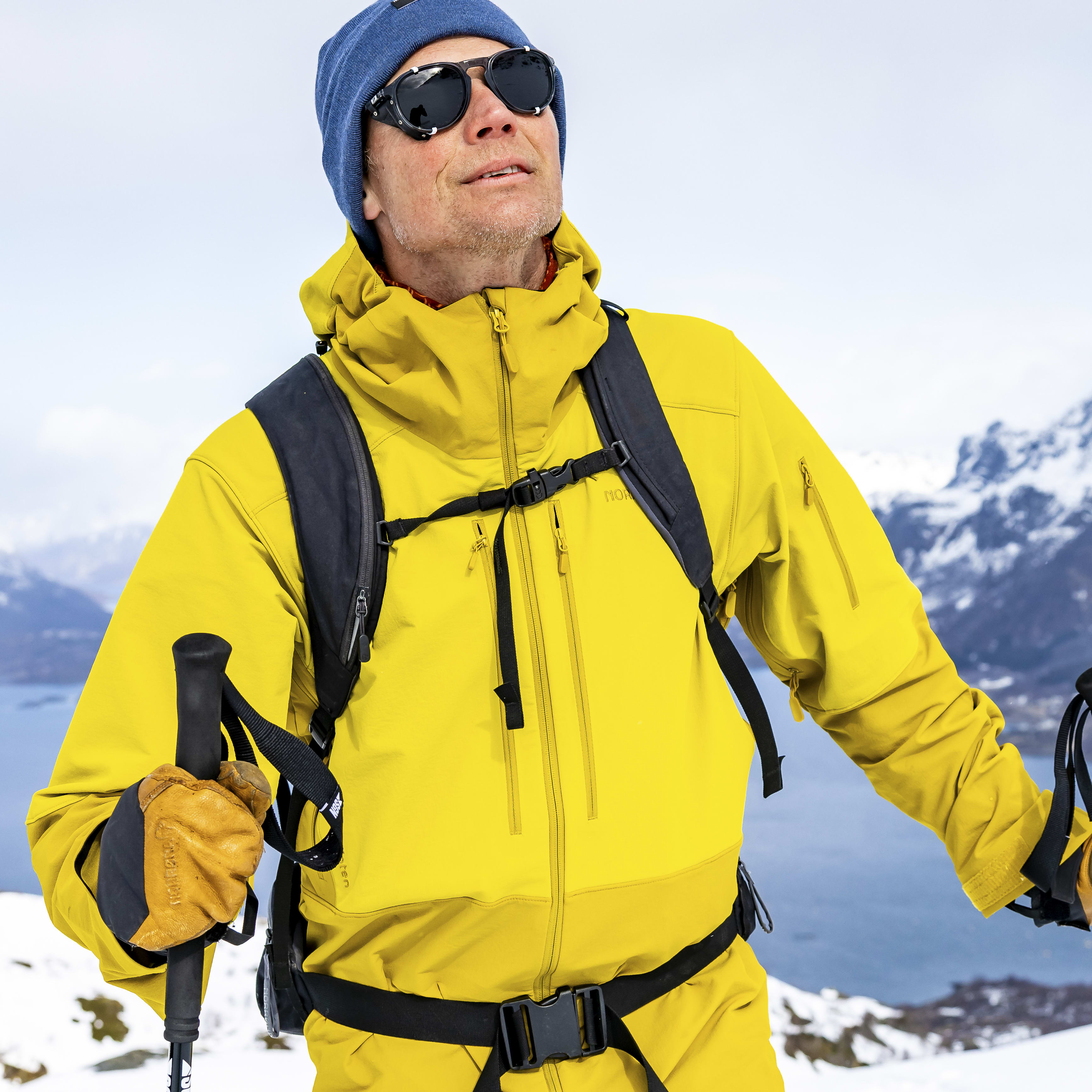 A man wearing a yellow Norrøna jacket and pants, a blue beanie, and sunglasses, stands in a snowy landscape holding ski poles.