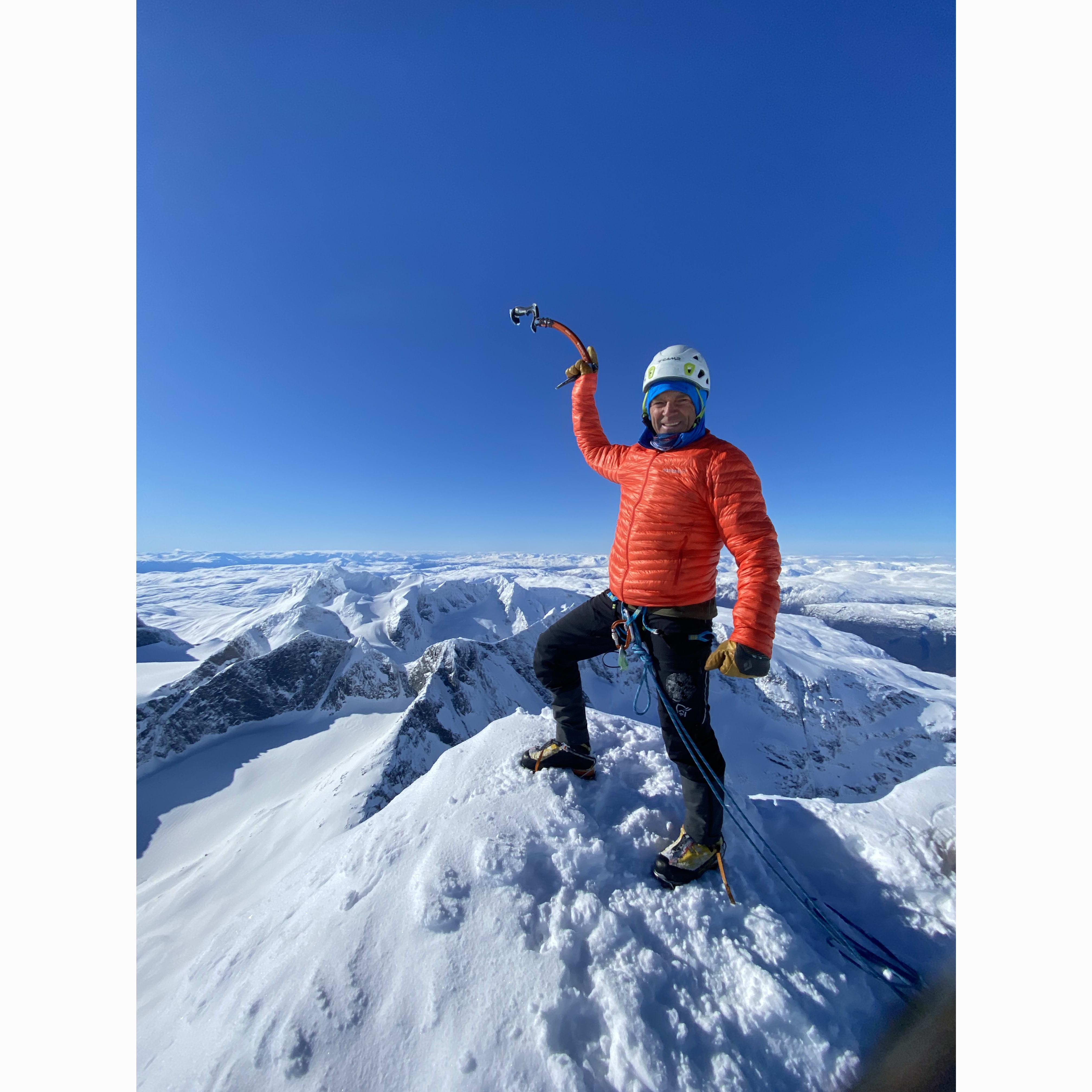A climber wearing a Norrøna jacket stands on a snowy mountain peak, holding an ice axe.  The sky is blue and clear. 
