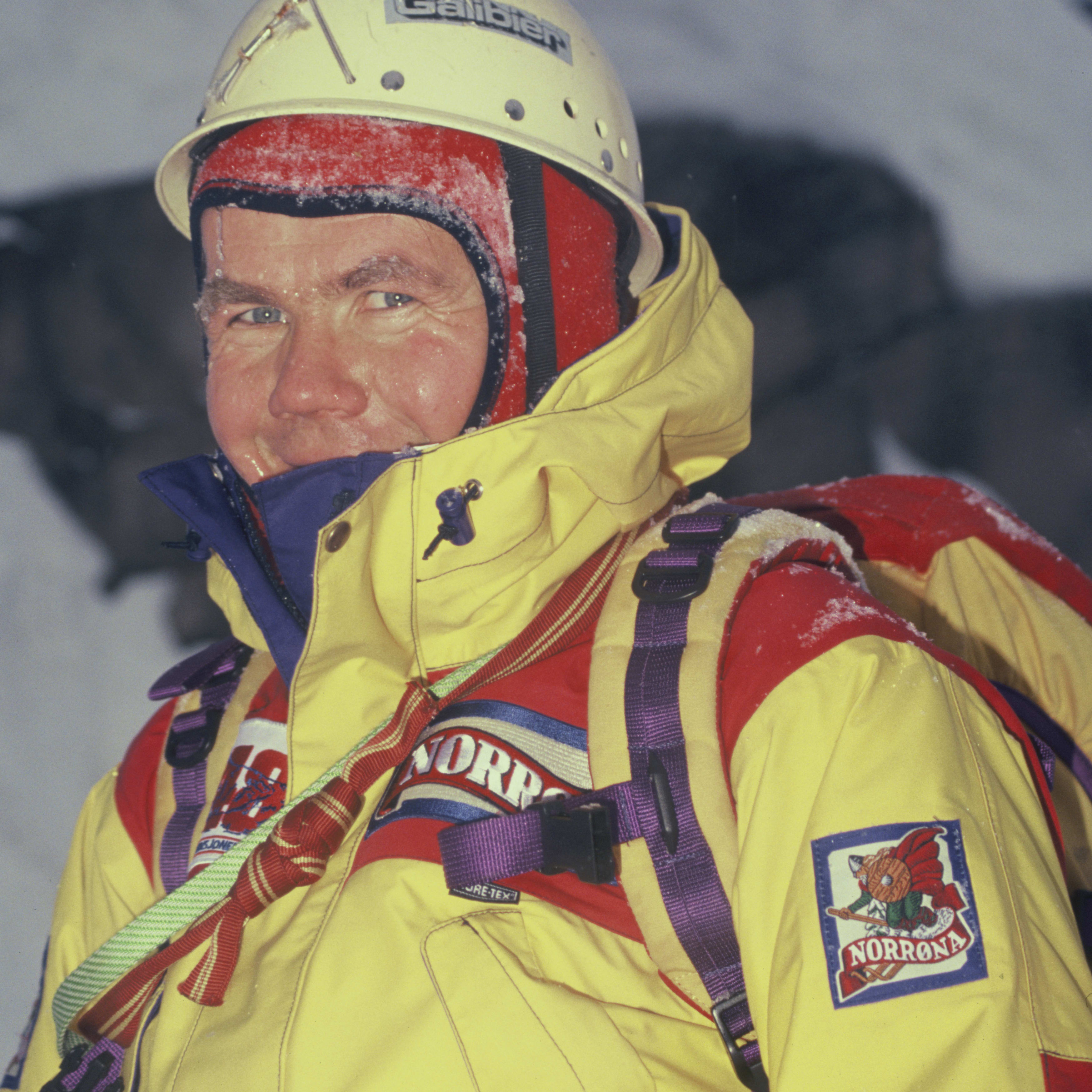 A man in a yellow, red, and blue jacket with a purple backpack, a white helmet with the words "Galibier" on it, and a patch on his jacket that says "Norrøna" is smiling into the camera. The man appears to be on a mountain and his face is covered in snow.