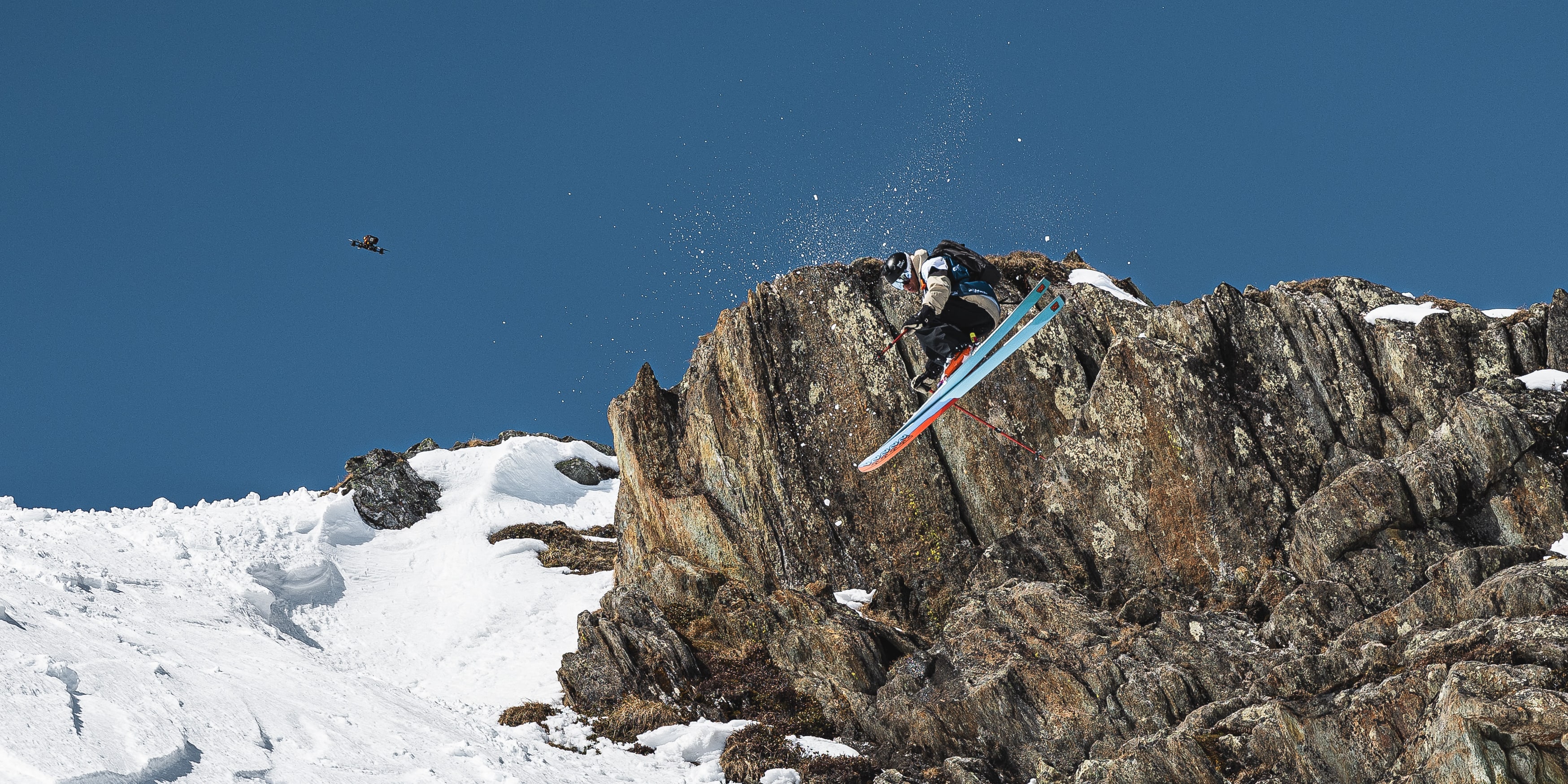 A skier wearing Norrøna clothing is airborne off a rocky cliff face on a sunny, snowy mountain. Another skier is visible in the background.