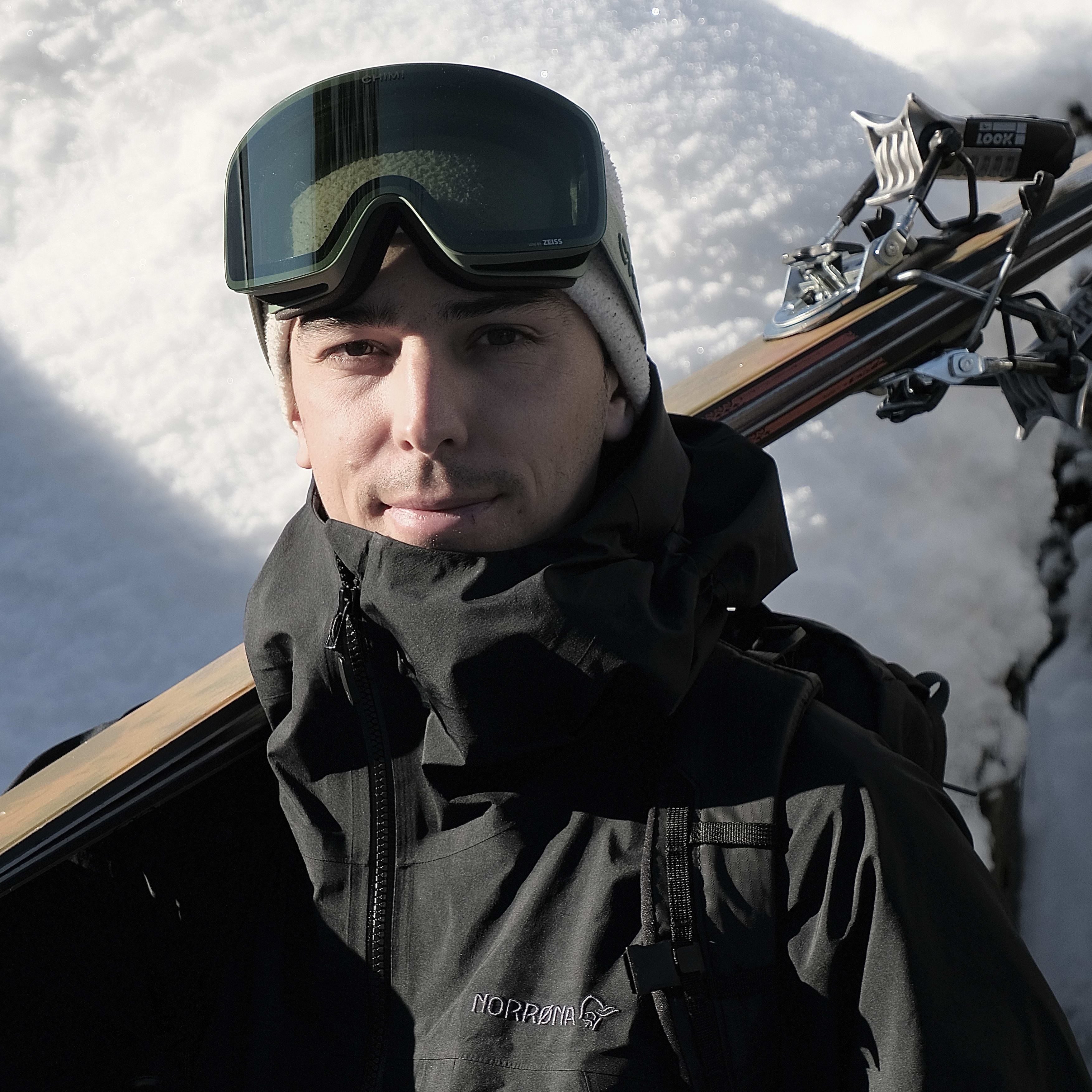 Man wearing a black Norrøna jacket and ski goggles, standing in a snowy landscape with a ski on his shoulder.  
