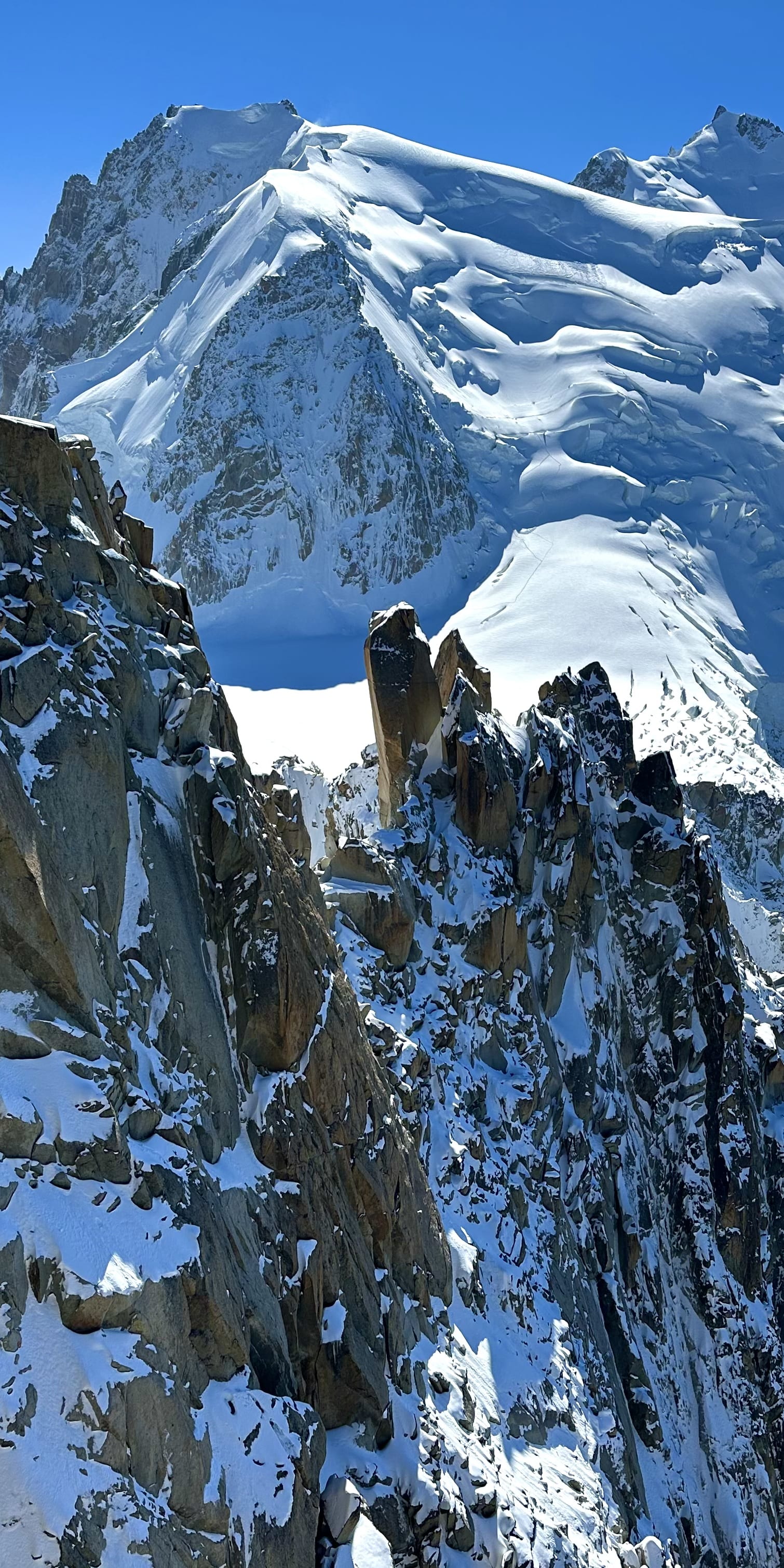 A panoramic view of snow-capped mountains. The foreground features a rocky cliff with patches of snow.
