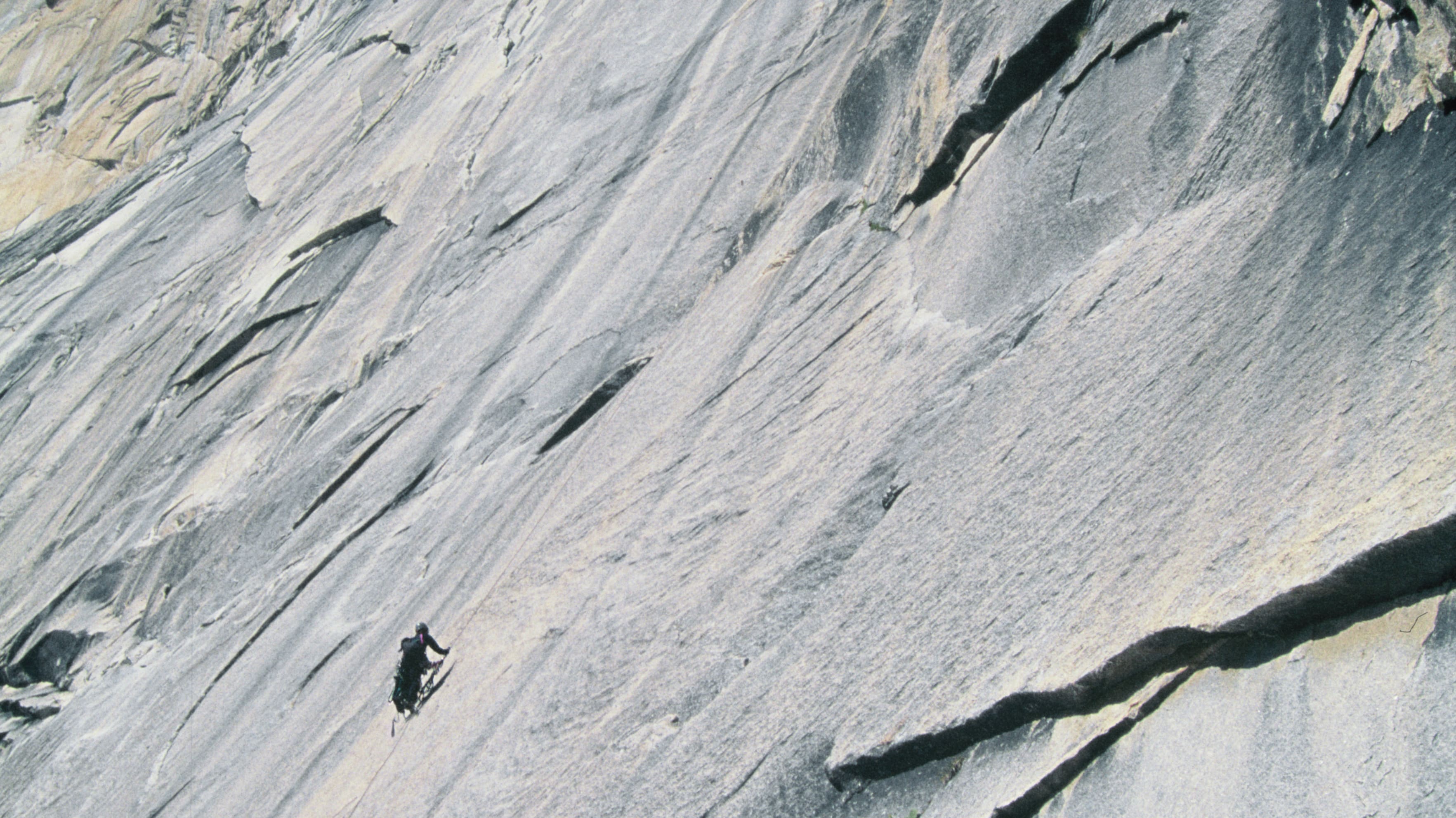 A climber, wearing Norrøna apparel, ascends a vast, gray granite rock face under a blue sky with a few clouds.
