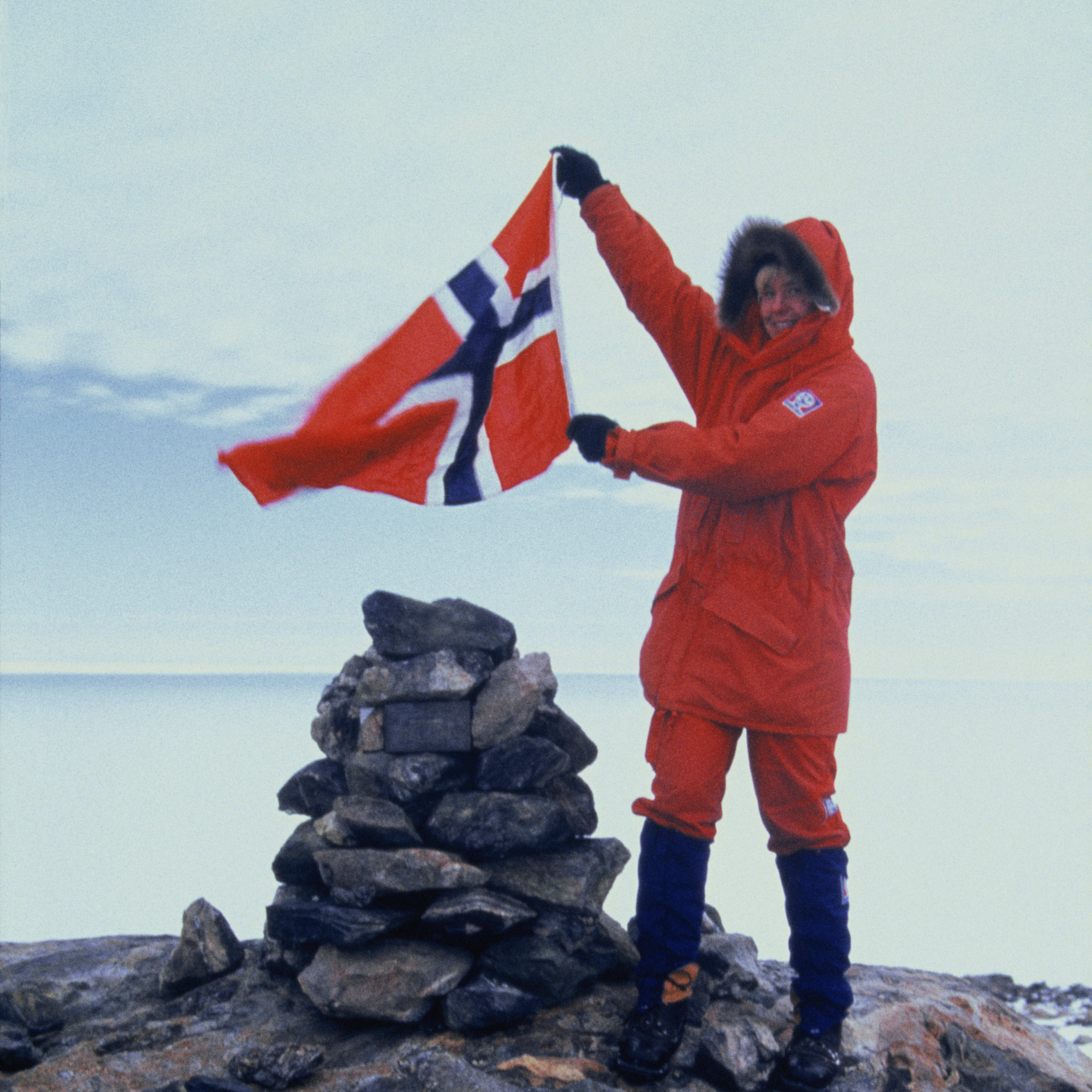A person in a red jacket and blue pants stands on rocky terrain holding a waving Norwegian flag.  They stand near a small cairn of rocks.