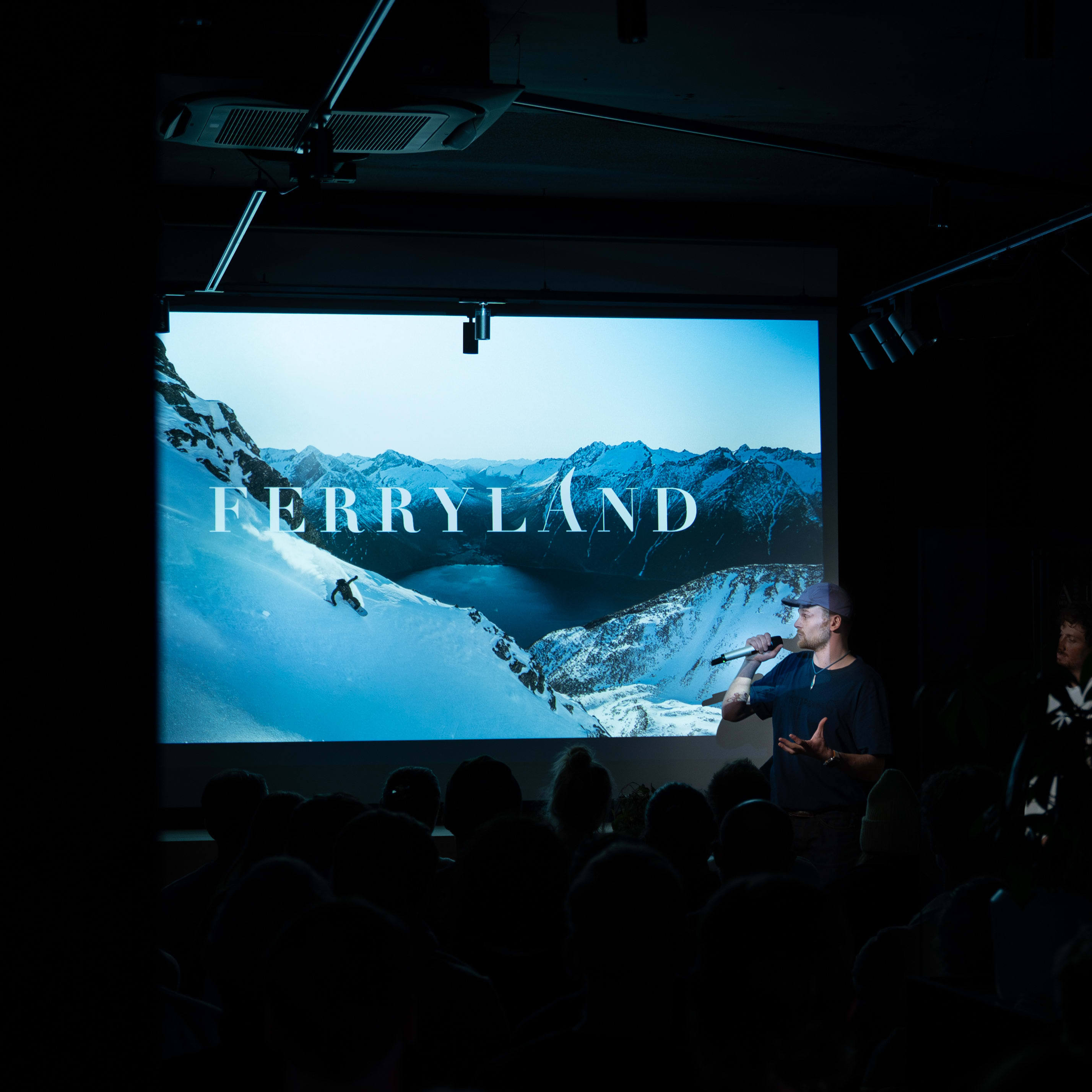 Presenter in Norrøna clothing speaks to an audience in front of a "FERRYLAND" movie screen.