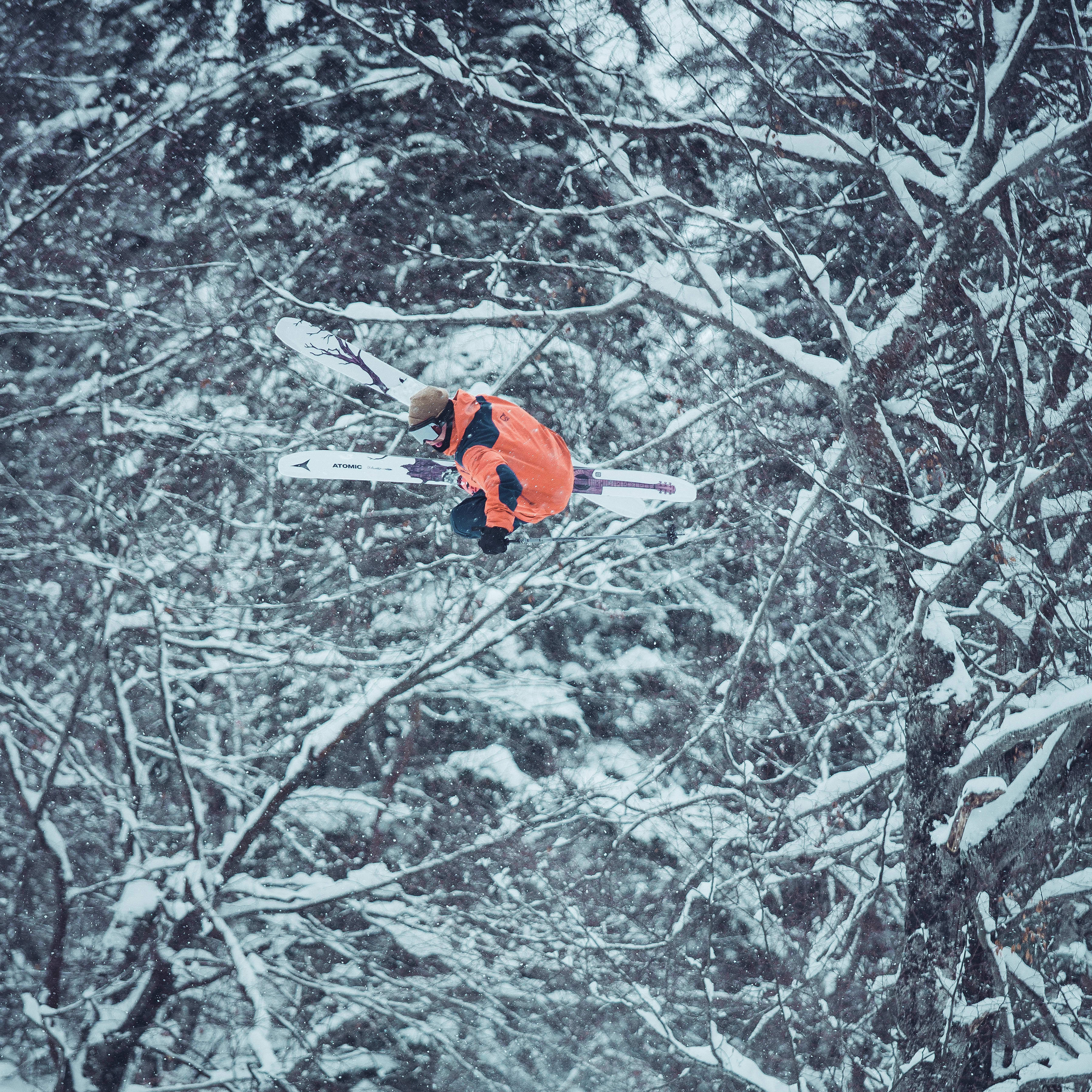 A skier in an orange Norrøna jacket performs a backflip in the air over a snowy slope and trees. 
