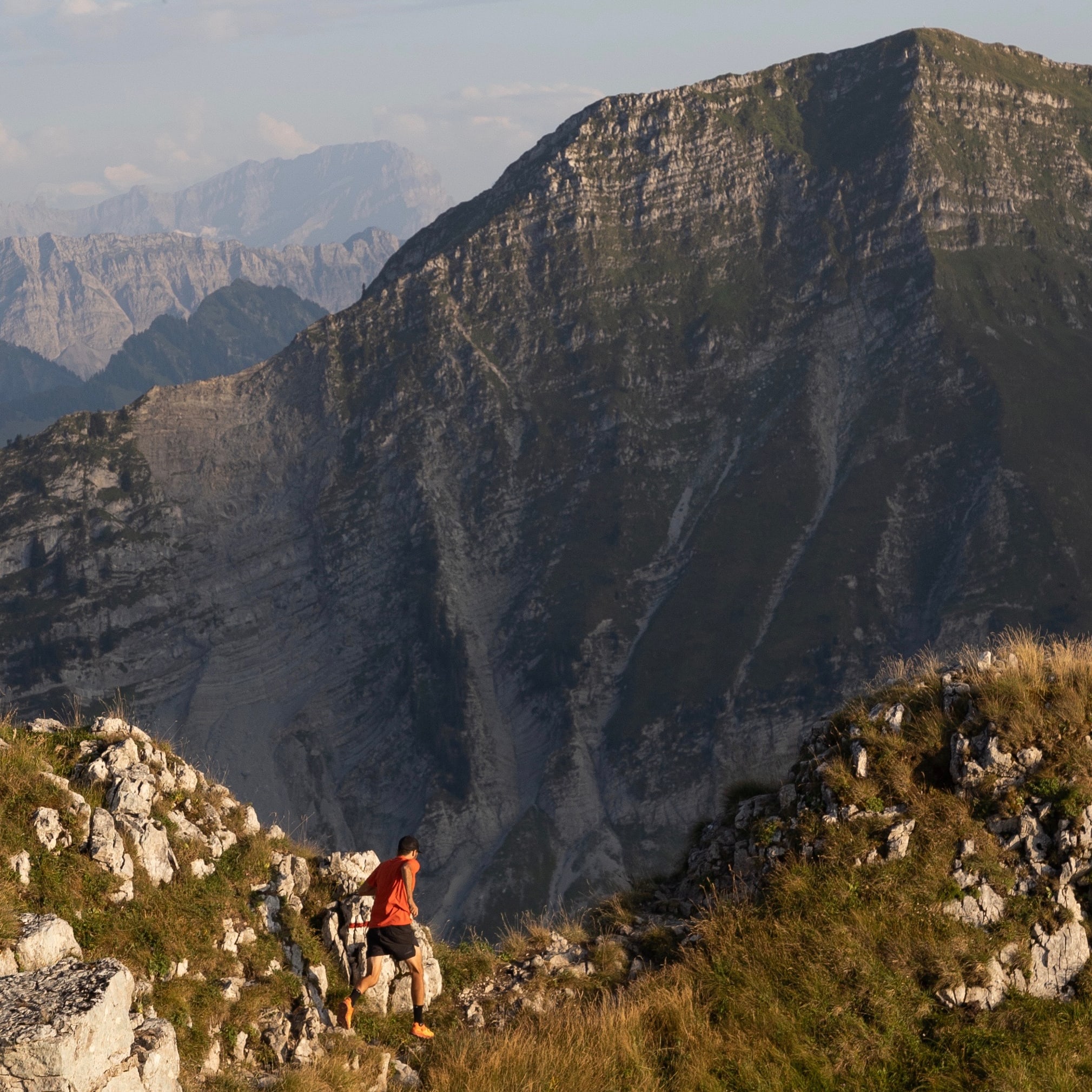 A person wearing a Norrøna shirt and shorts hikes on a rocky mountain ridge with a view of distant mountains. 
