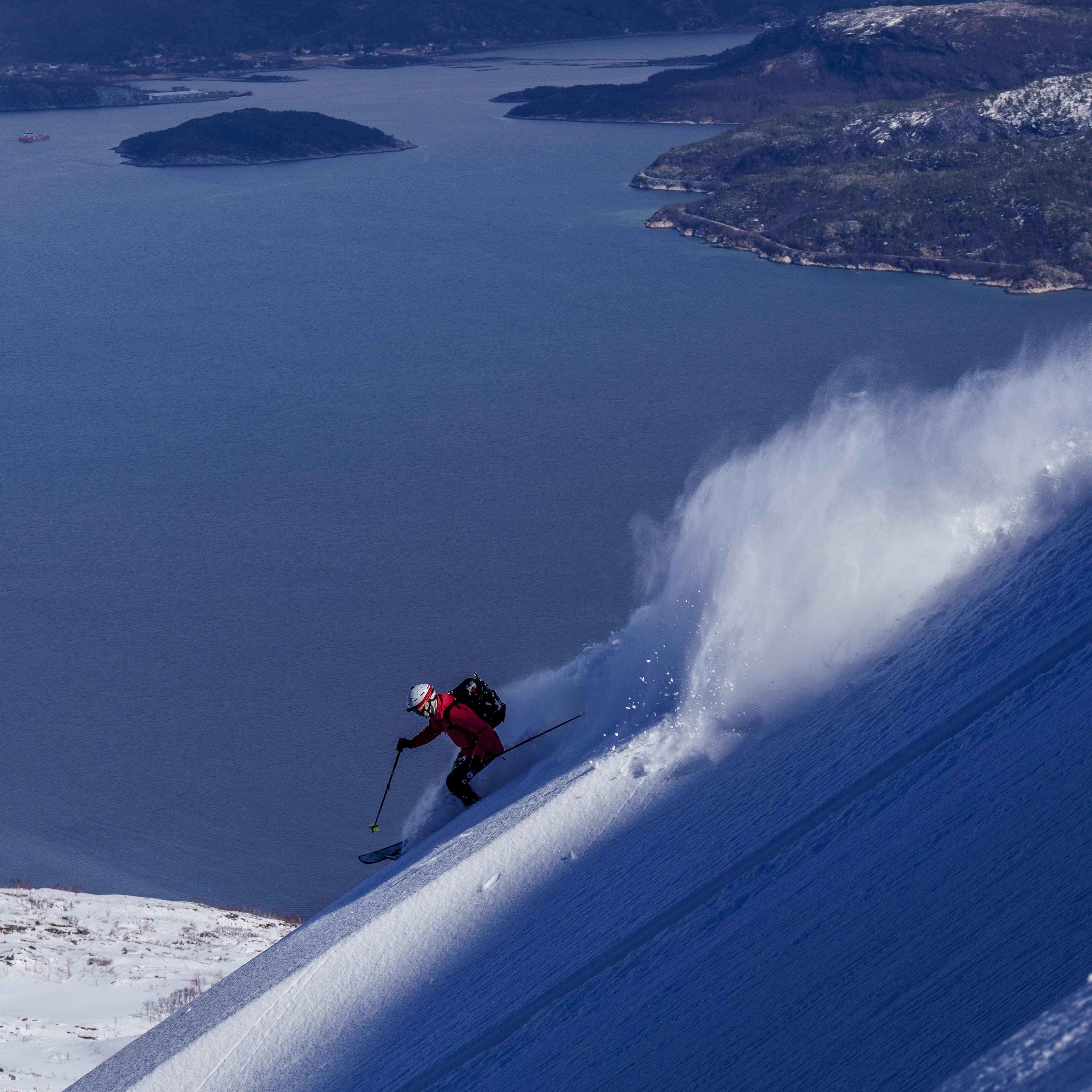 A skier in a red Norrøna jacket skis down a steep, snowy slope, kicking up powder. Blue water and islands fill the background.
