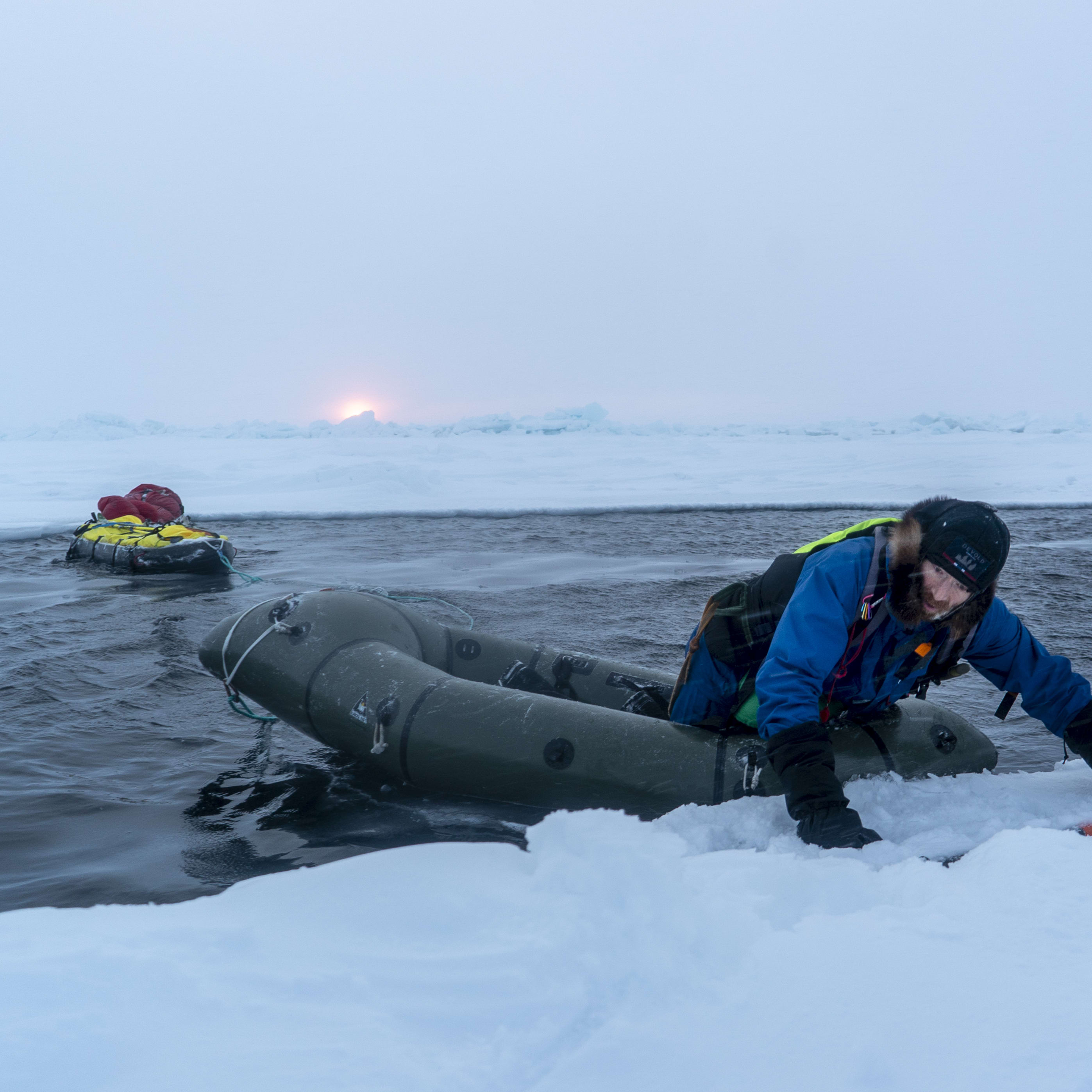 A person in Norrøna clothing exits an inflatable boat onto an ice floe. A second boat with supplies floats nearby. The sky is overcast with a low sun.