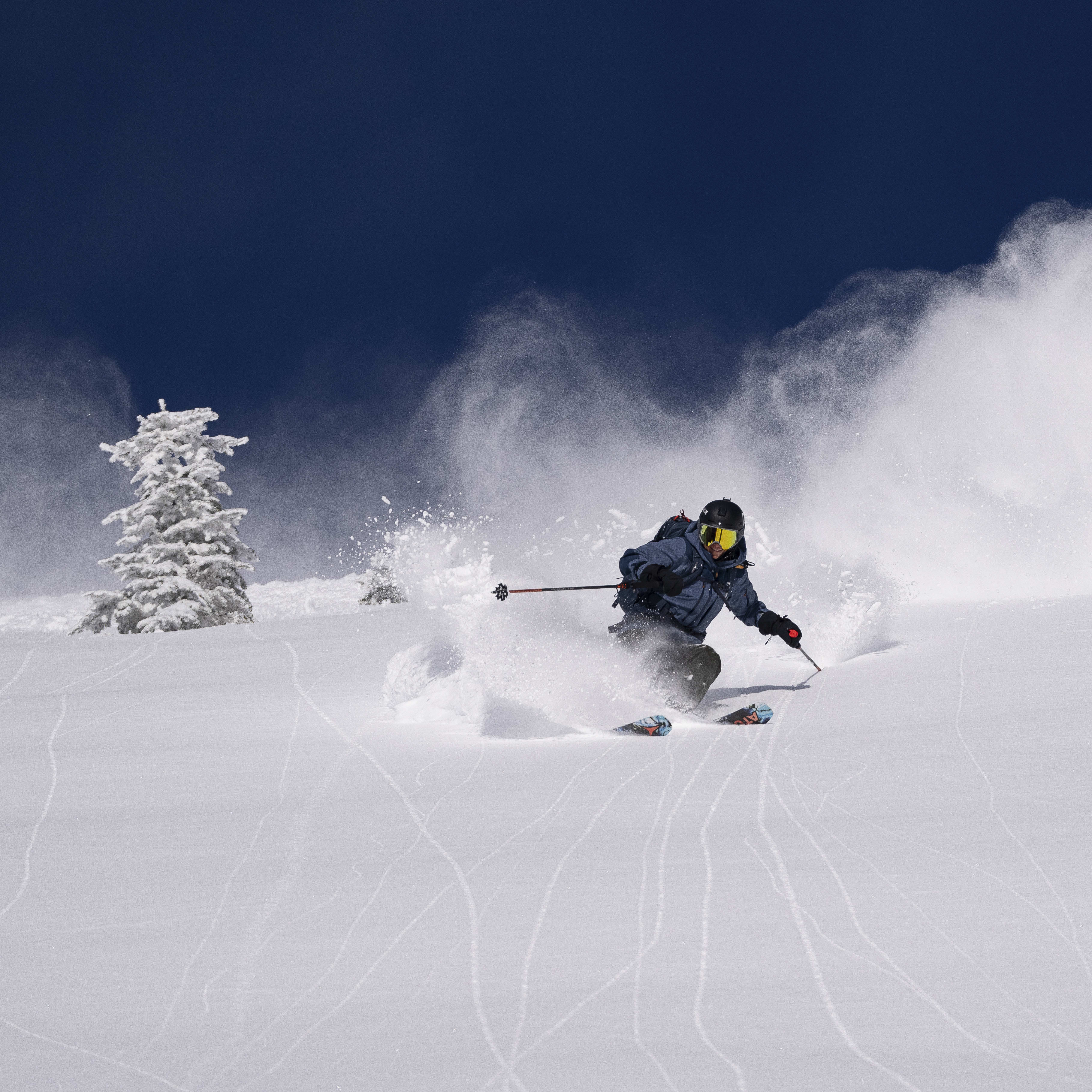 A skier wearing a Norrøna jacket and goggles makes a turn on a snowy slope, creating a spray of powder.