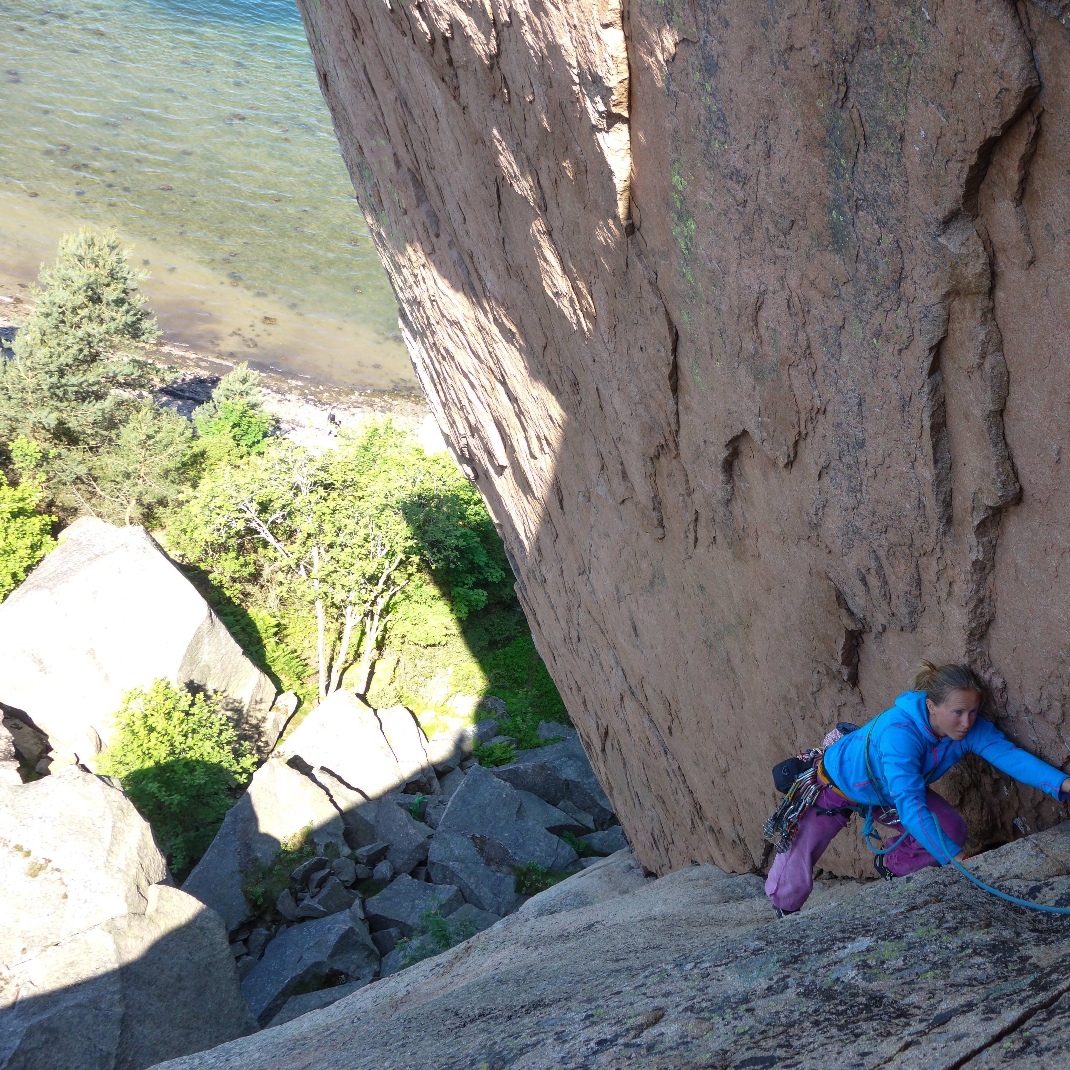 A woman rock climbs on a sunny day, secured by a rope. She wears a blue Norrøna jacket and purple pants. The ocean and foliage are visible in the background.
