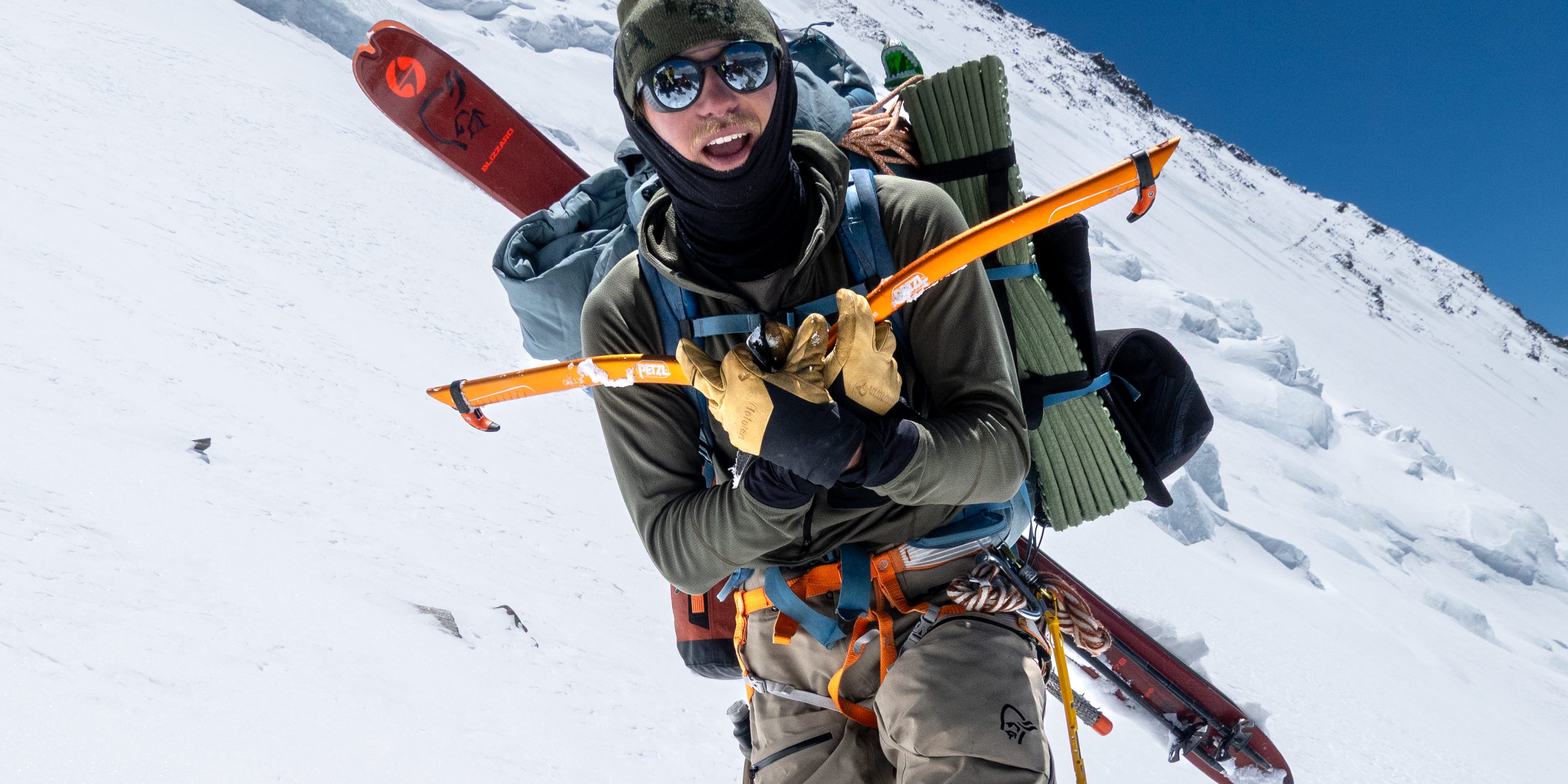A climber in Norrøna pants ascending a snowy mountain. He carries skis and ice axes on his backpack. Blue sky is visible.