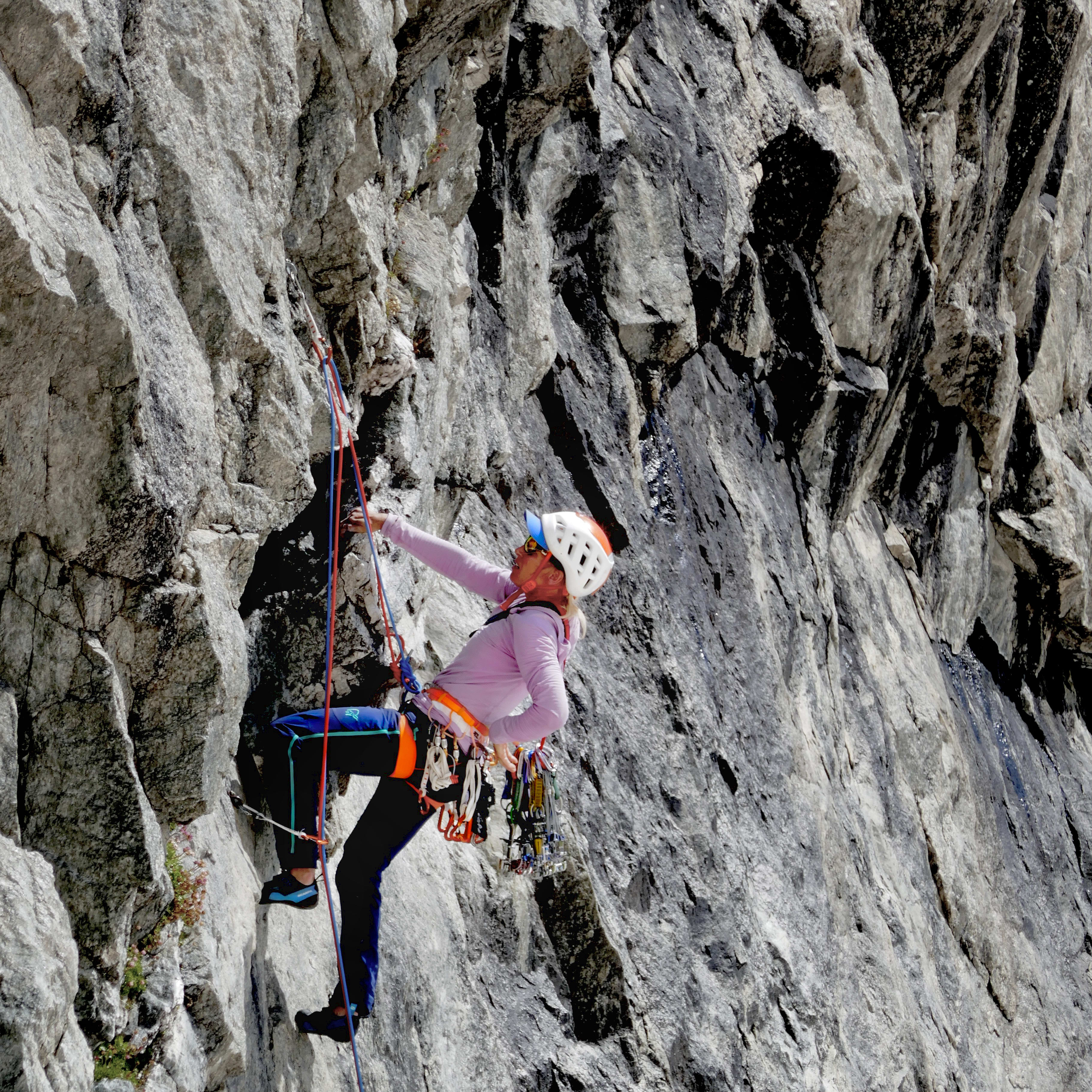 A climber in Norrøna pants scales a grey rock face, secured by ropes and gear, and wearing a helmet.