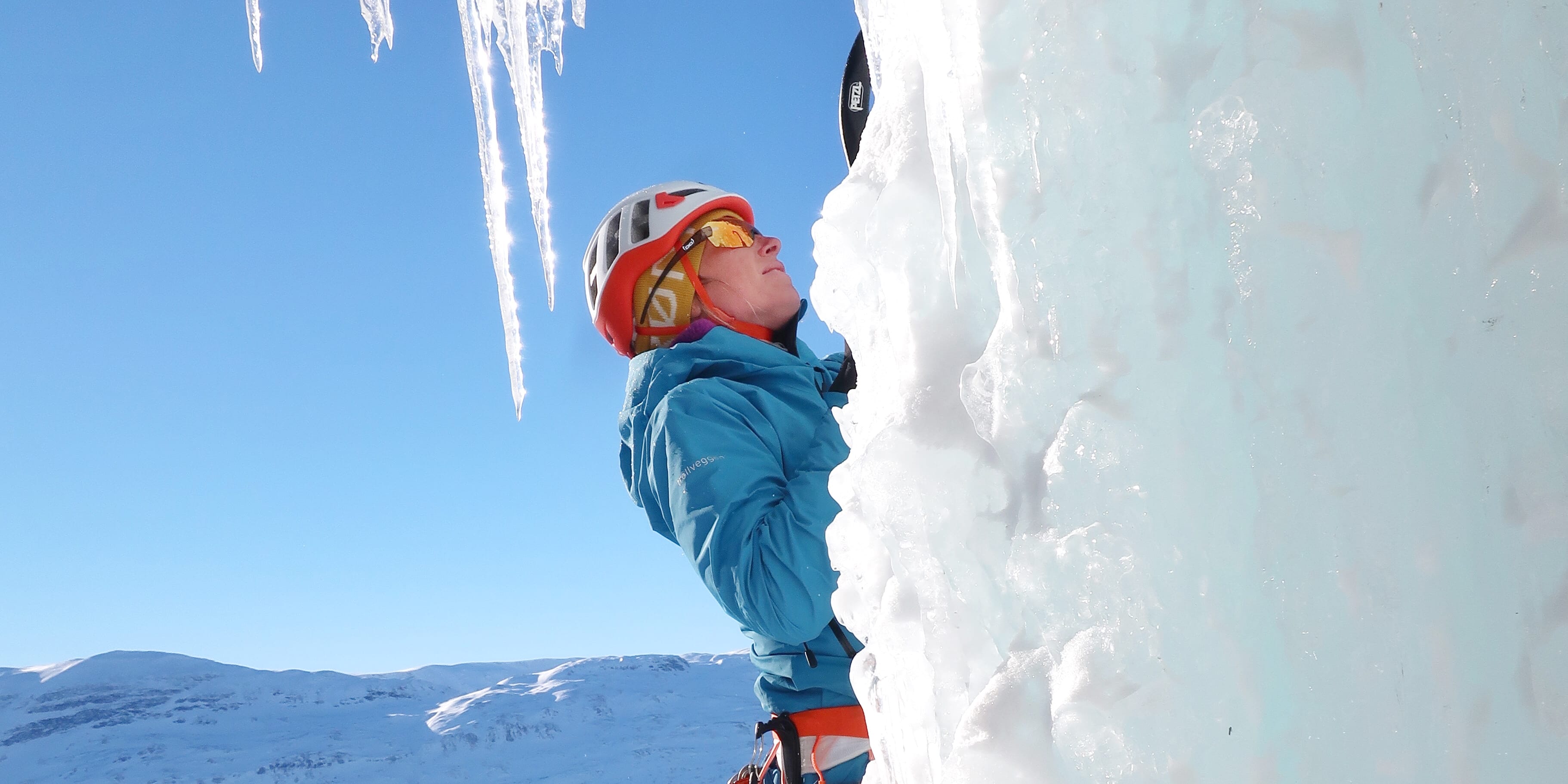 An ice climber with a helmet and Norrøna jacket ascends a frozen wall with icicles. Snow-covered mountains are in the background.
