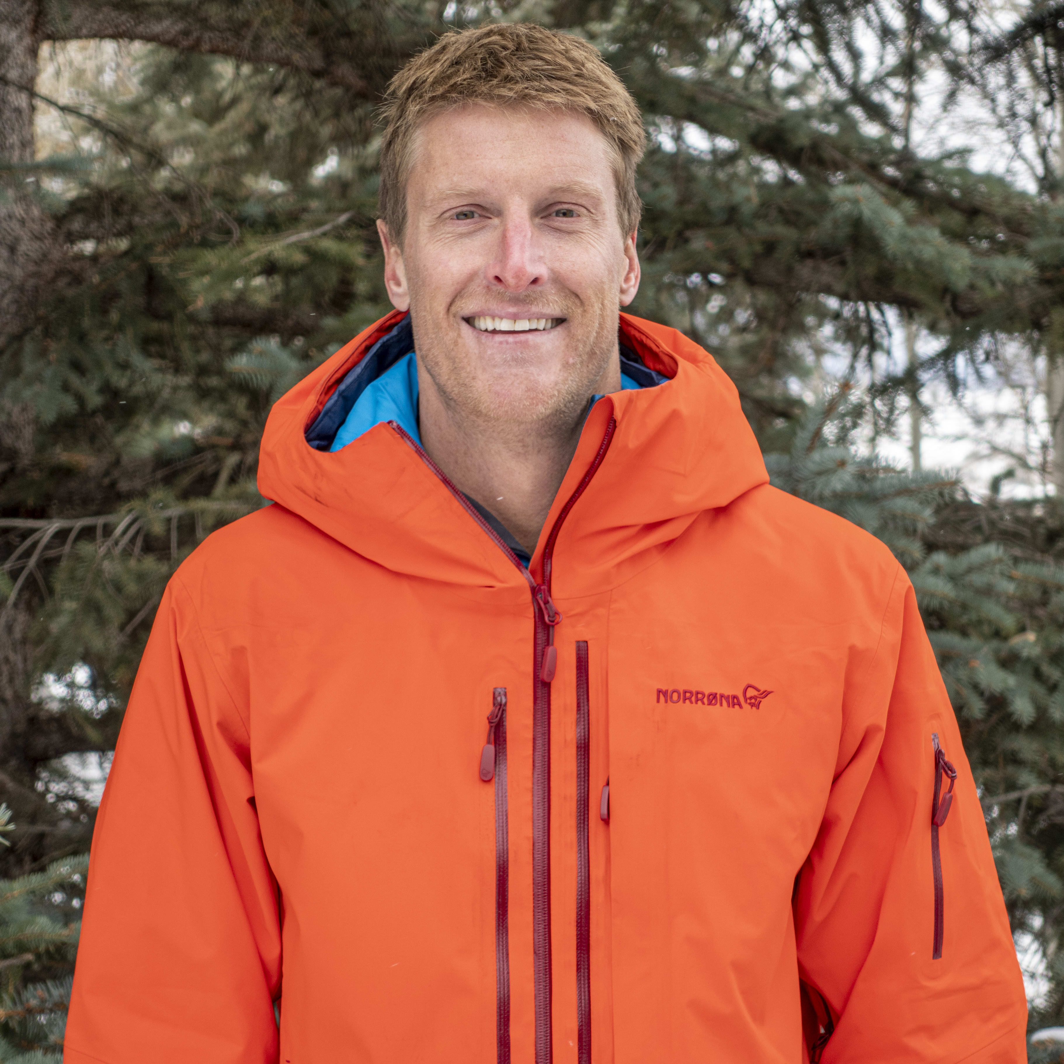 A smiling man in a bright orange Norrøna hooded jacket stands in front of a pine tree.