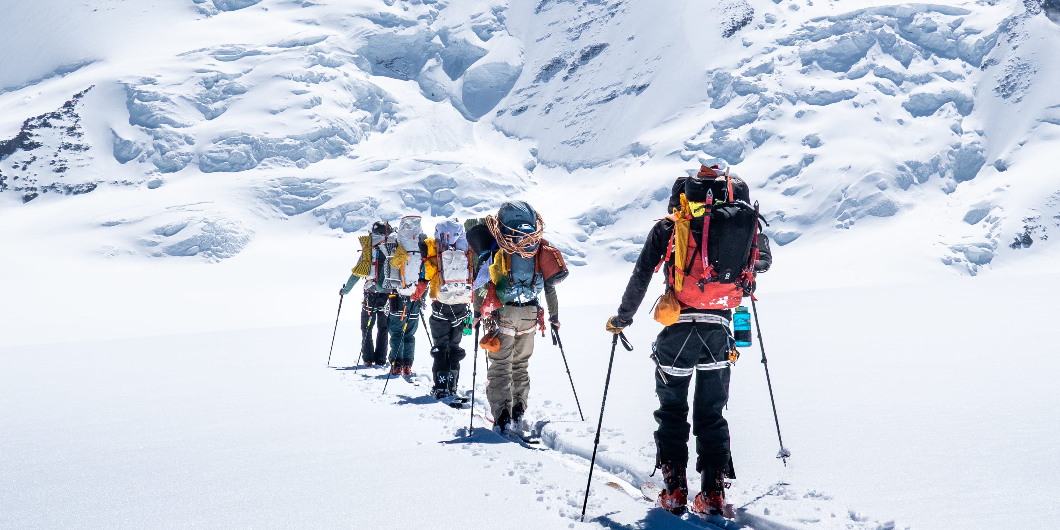 A group of mountaineers on skis, wearing Norrøna apparel and backpacks, traverse a snowy field towards a large, snow-covered mountain under a clear blue sky.