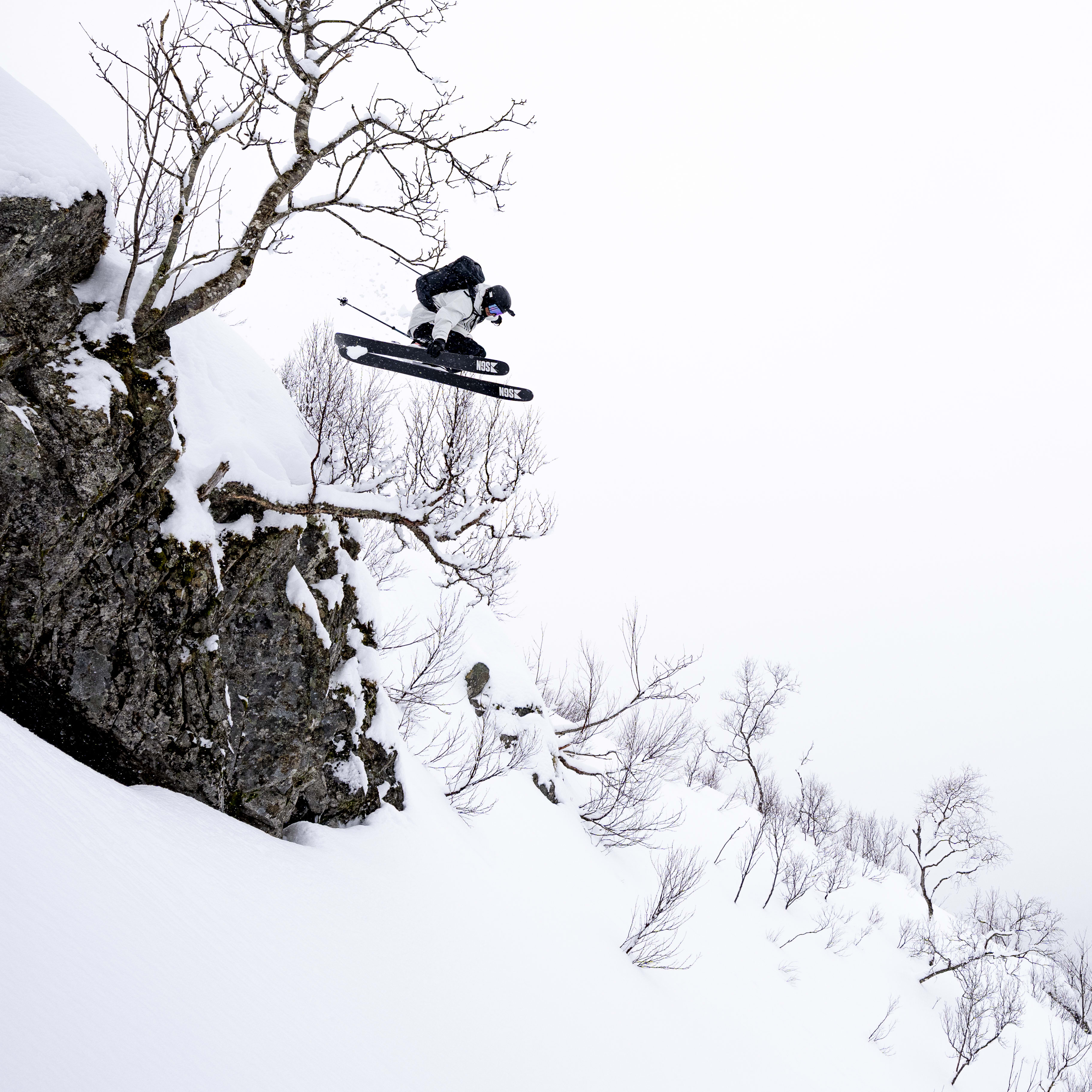 A skier wearing a Norrøna jacket jumps off a cliff in the snow. The skier's skis are visible. The background is a snowy mountain range.