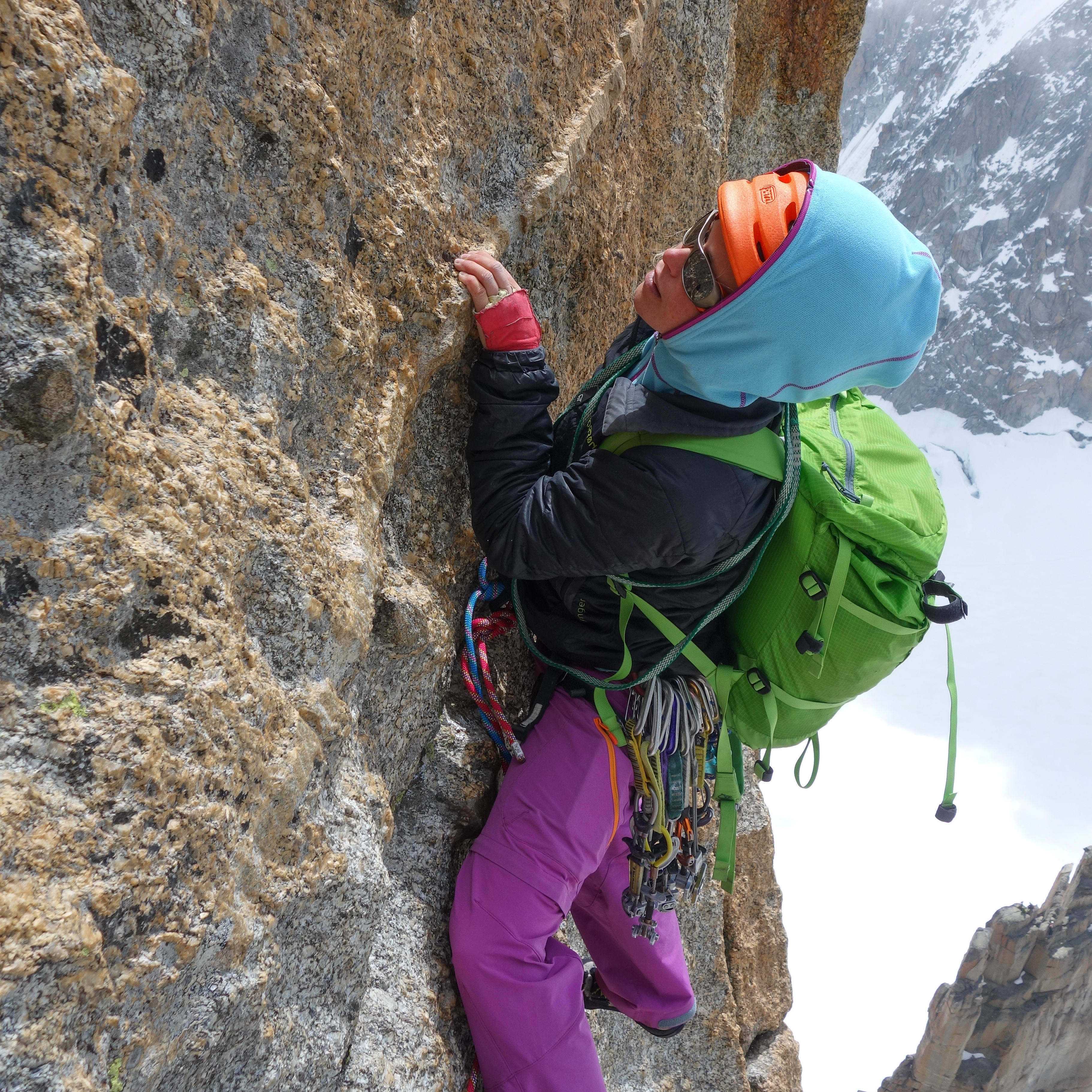 A climber in Norrøna pants ascends a rock face. They have a green backpack and are surrounded by climbing gear, with snow-covered mountains in the background.
