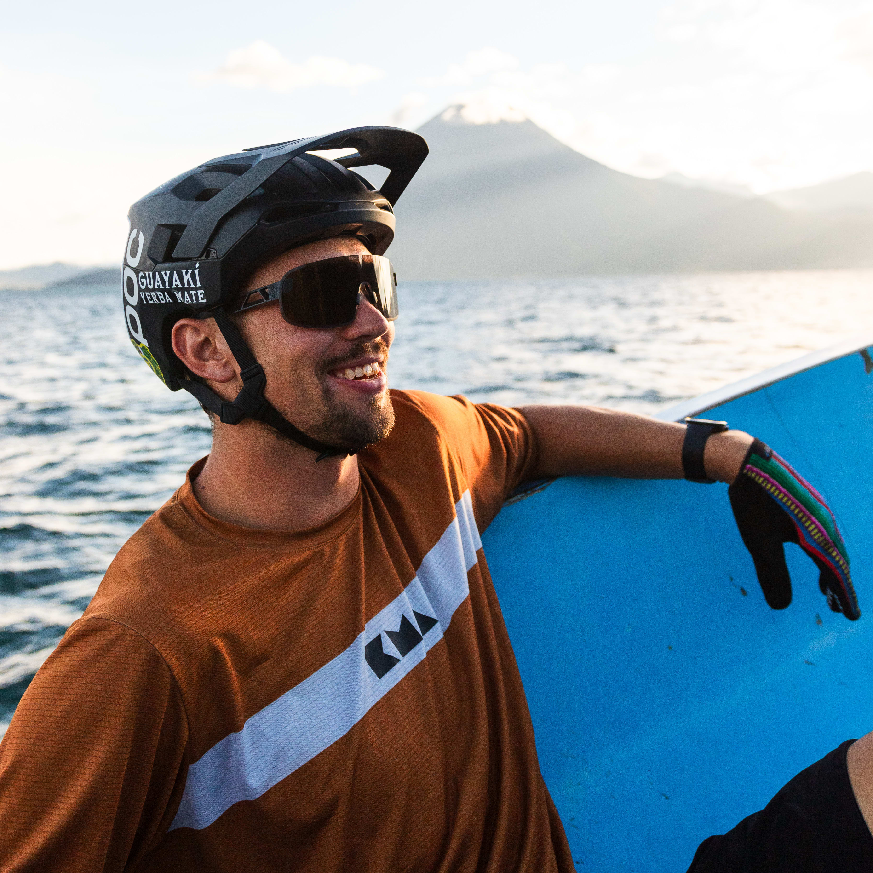 A smiling person on a blue boat, wearing sunglasses, helmet, and a brown Norrøna shirt. A mountain is in the background.