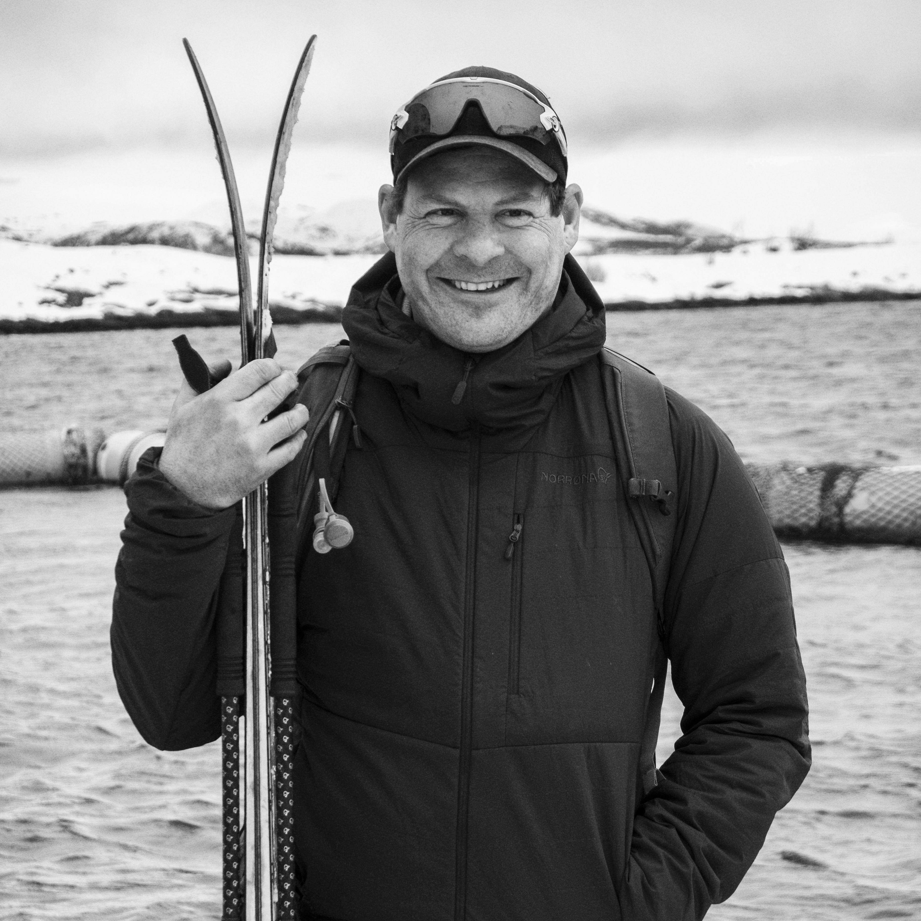 A man wearing a black Norrøna jacket and a baseball cap holds two skis in his hand, standing in front of a body of water. 
