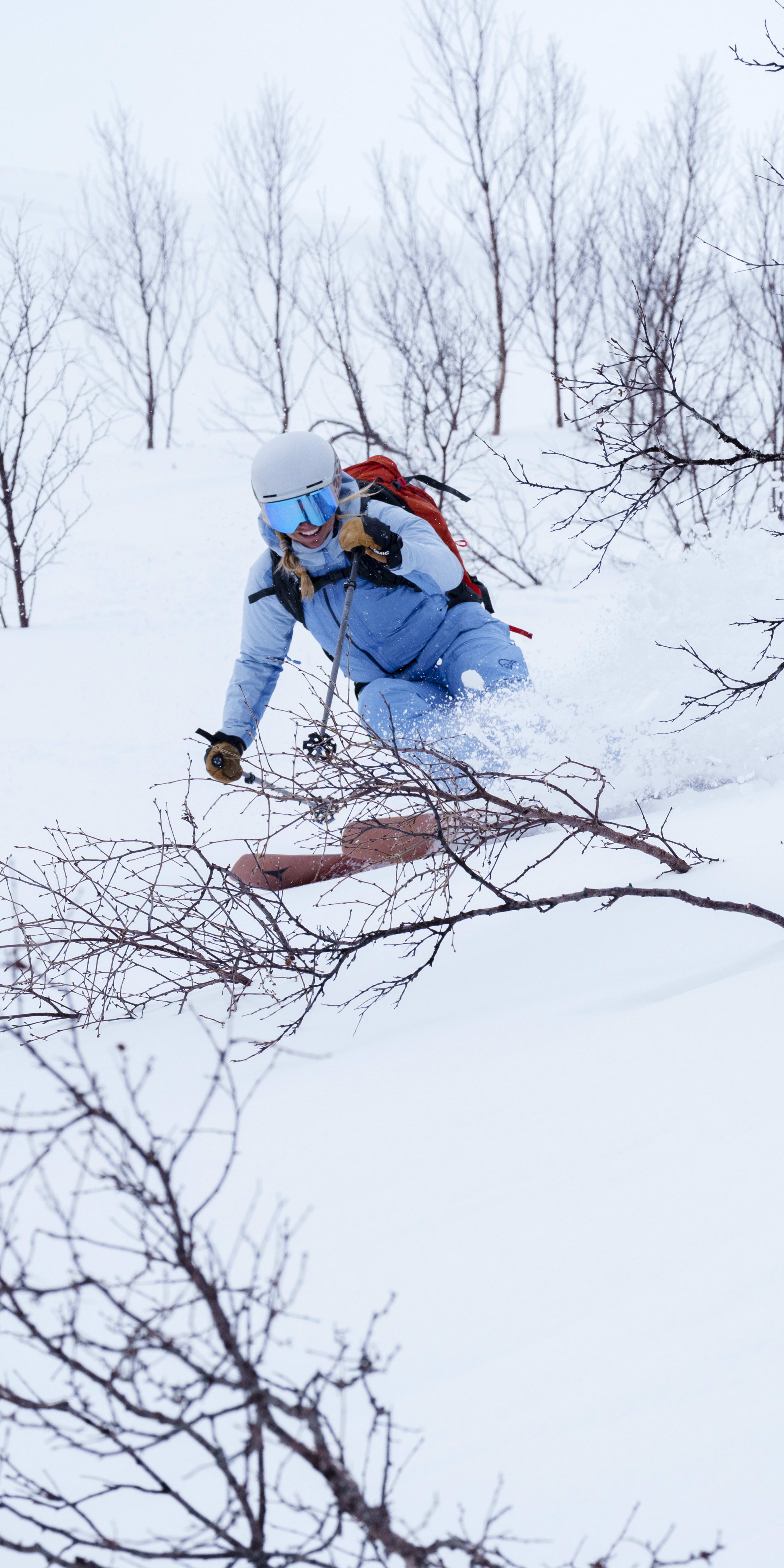 A skier in a blue Norrøna suit skis through deep snow on a cloudy day. The skier wears a helmet, goggles and a backpack, and is surrounded by trees.