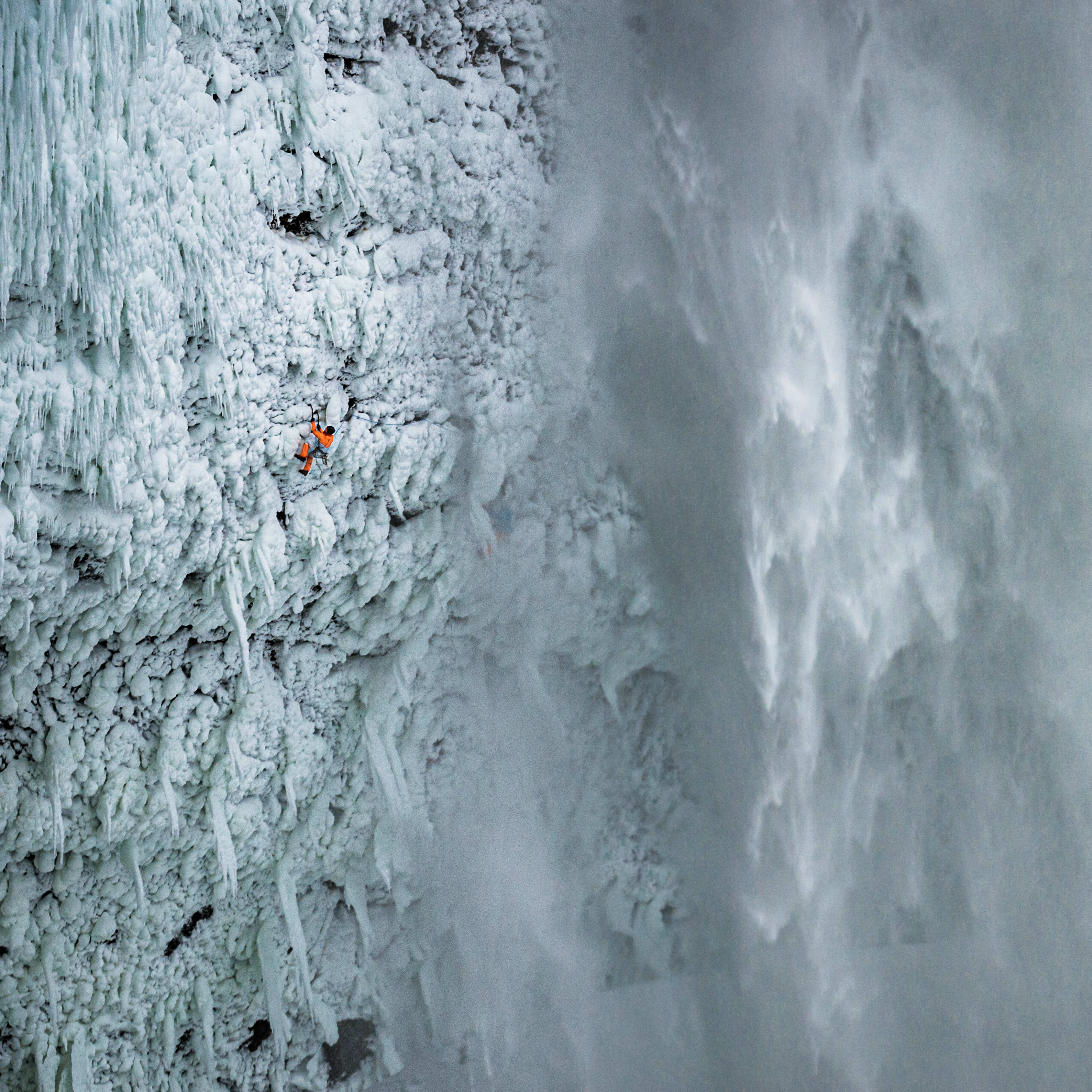 A climber, wearing Norrøna gear, scales an icy waterfall. The water is frozen and the climber is small in the vast landscape.