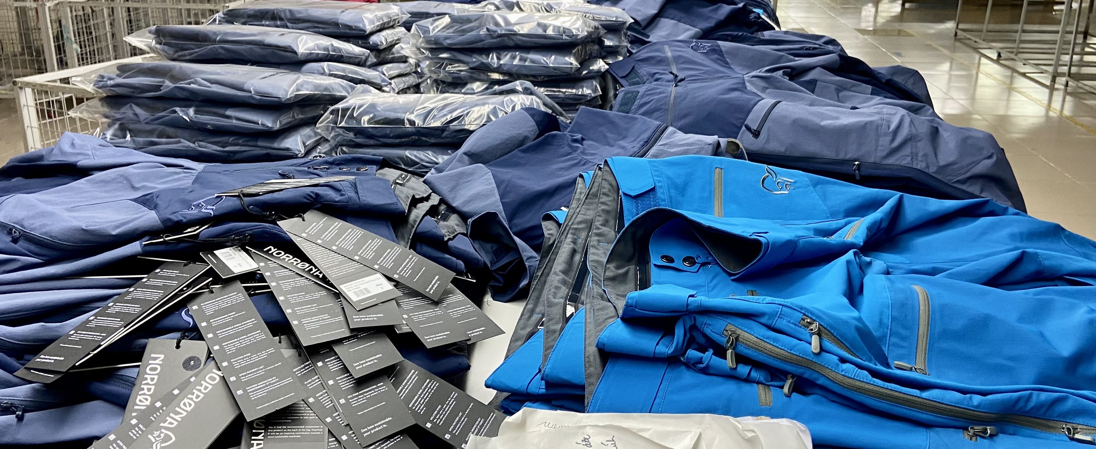 A stack of folded Norrøna clothing, mostly in blue and black, with several tags visible.  A worker stands near the pile in a factory setting. 
