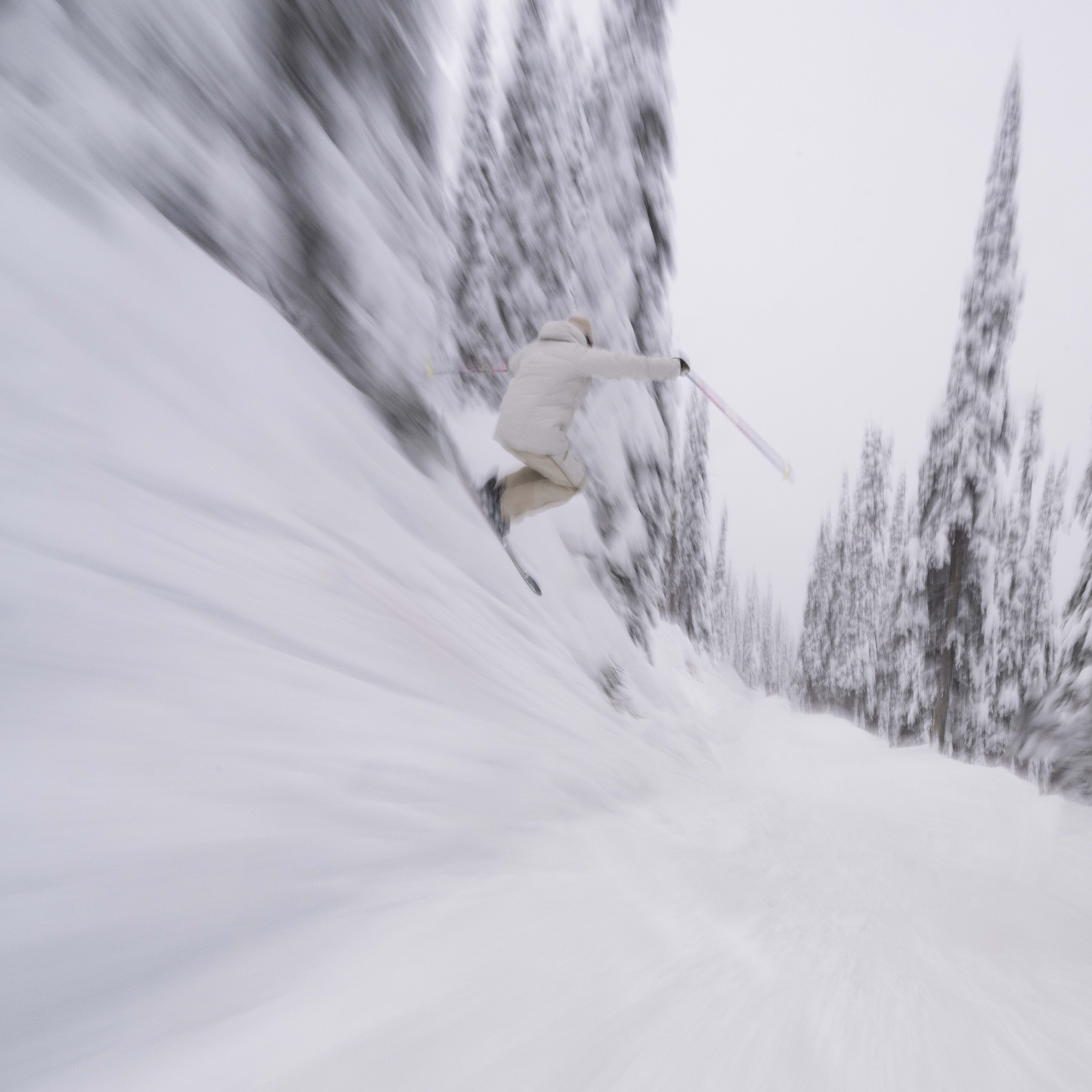 A skier in a Norrøna jacket speeds down a snowy slope. The trees lining the path are blurred due to motion.