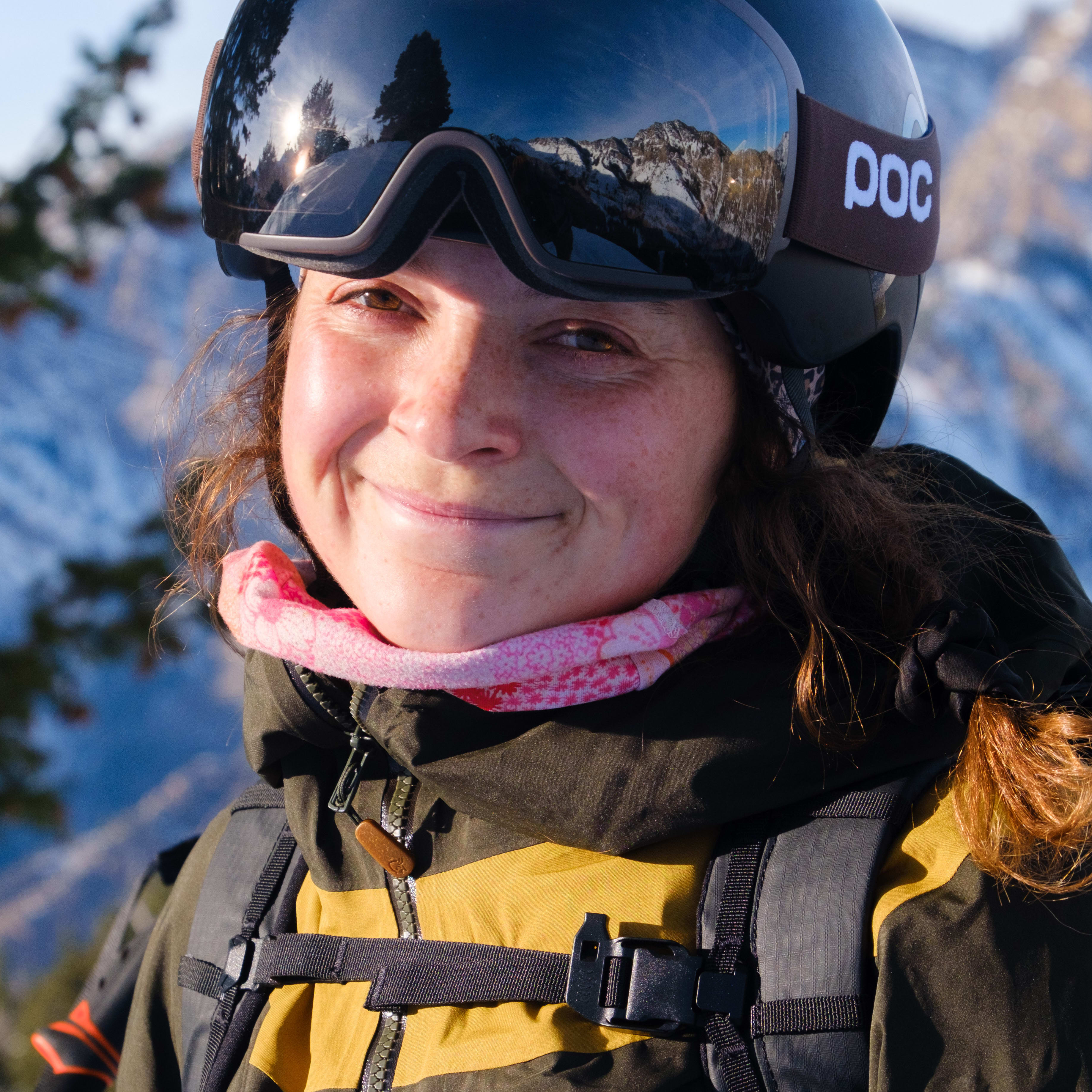 A woman wearing a black helmet with a white POC logo and Norrøna clothing smiles into the camera.  She is wearing a pink patterned neck gaiter, and has a black backpack with a yellow strap.