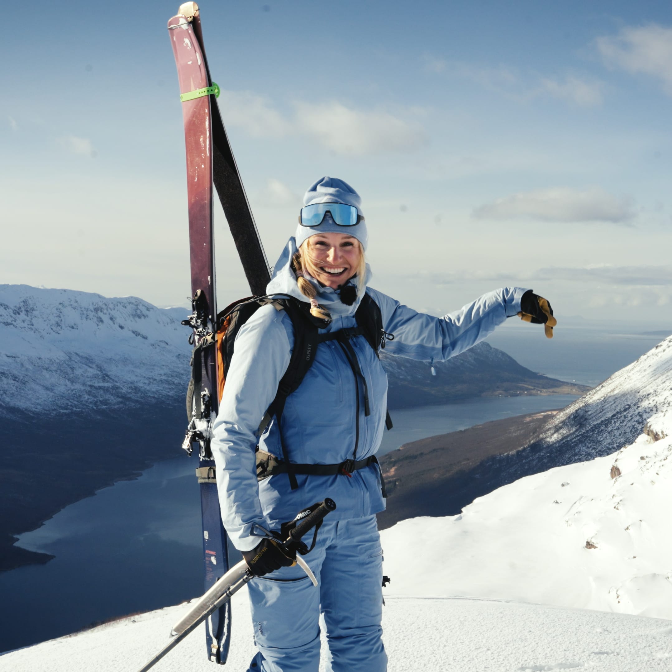 Smiling woman in blue Norrøna ski gear on a snowy mountain peak overlooking a fjord.
