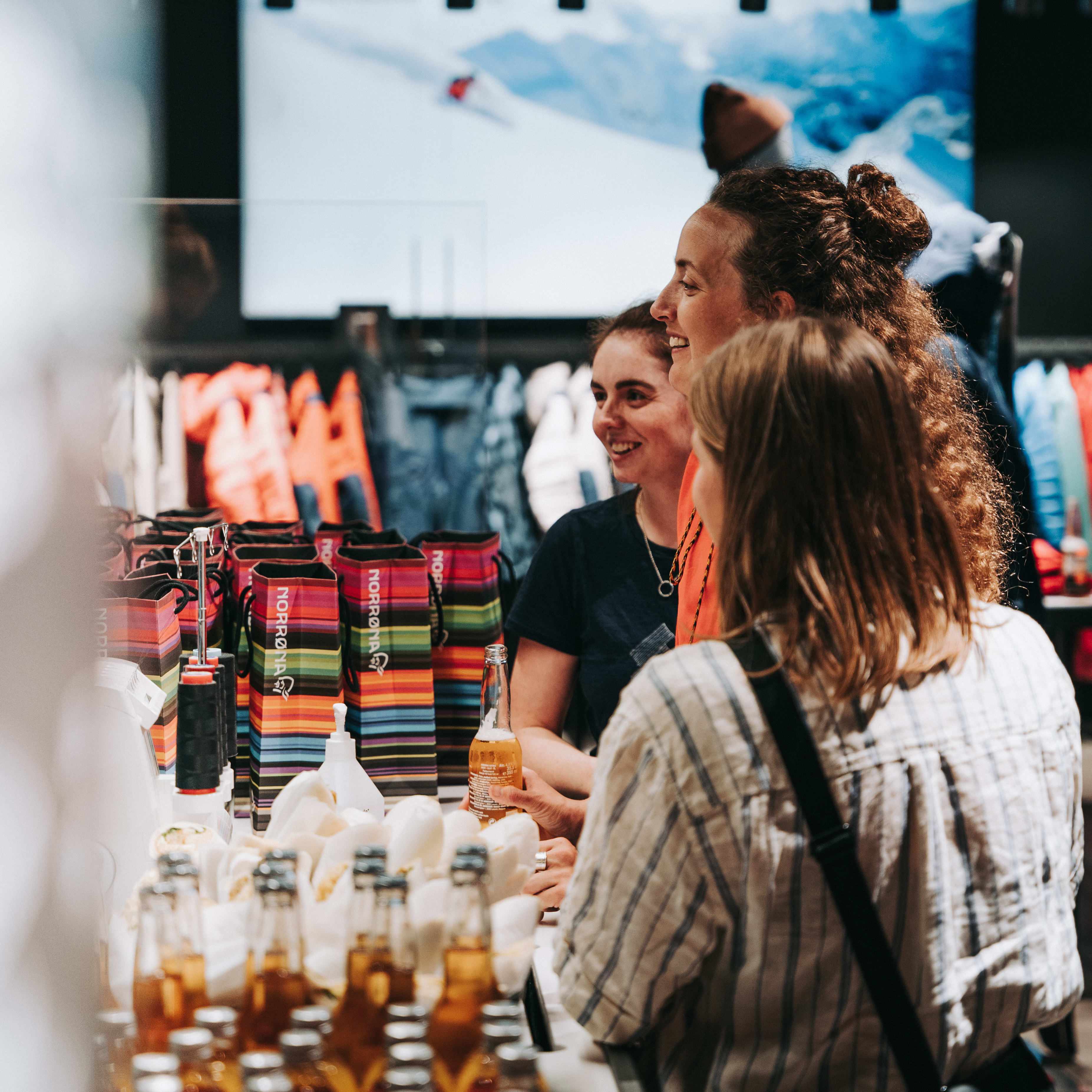 Three women smiling in a store. A row of Norrøna shopping bags are on the counter. One woman holds a bottle of beer.