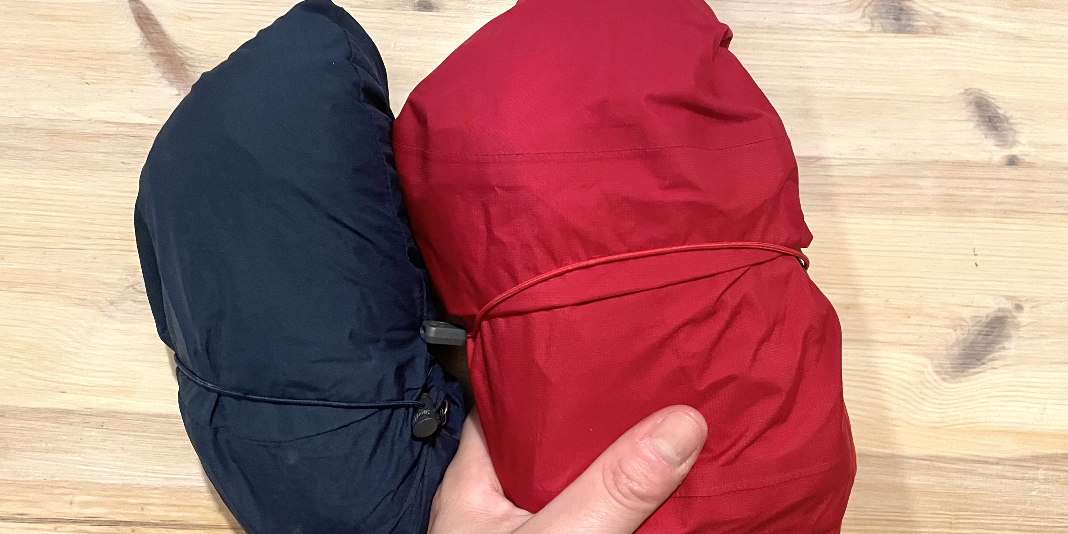 A hand holds a red Norrøna jacket, folded and secured with a red string. Beside it lies a folded blue Norrøna jacket on a wooden surface.