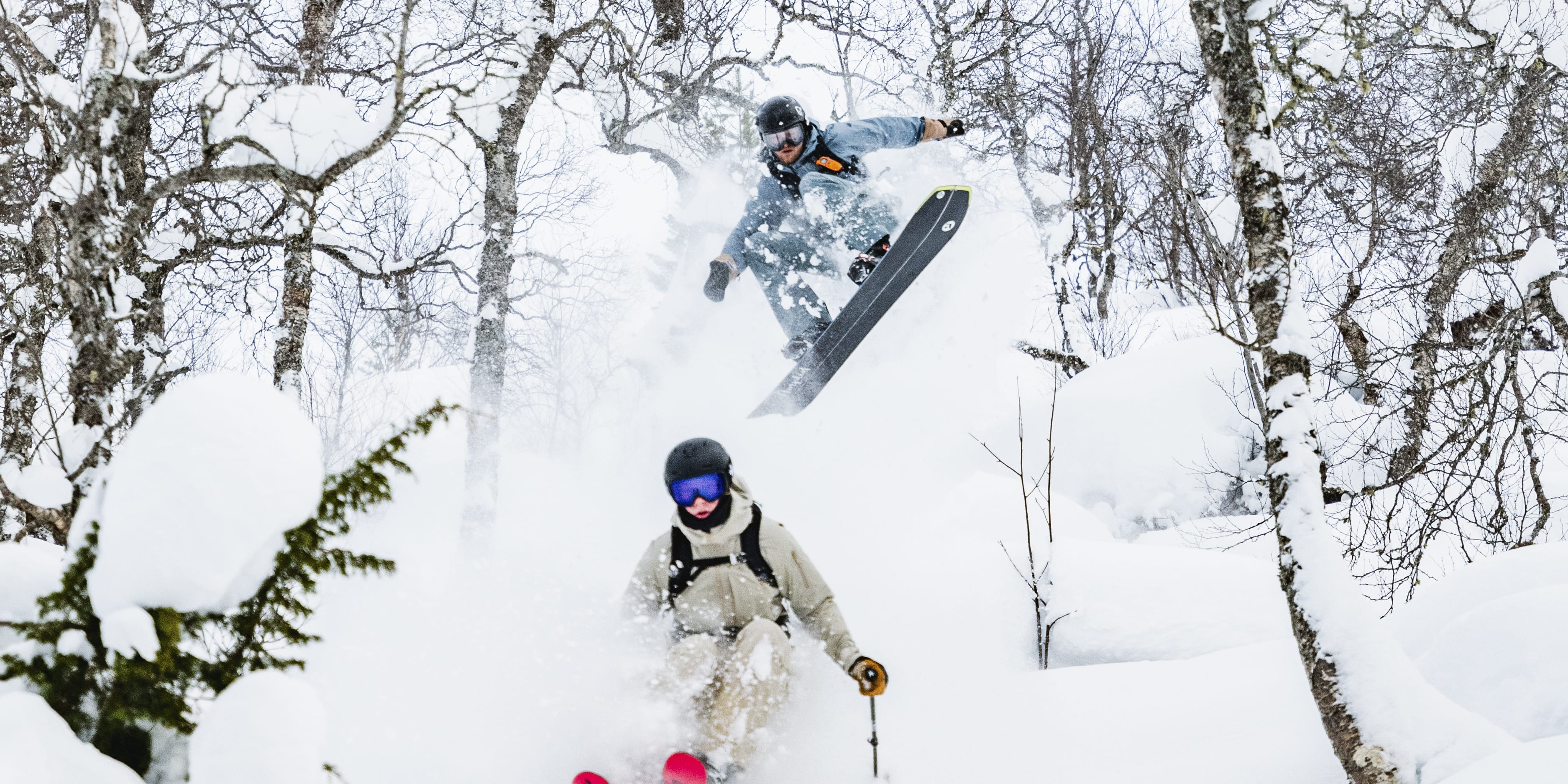 Two skiers in Norrøna gear ski down a snowy slope through a snowy forest. 
