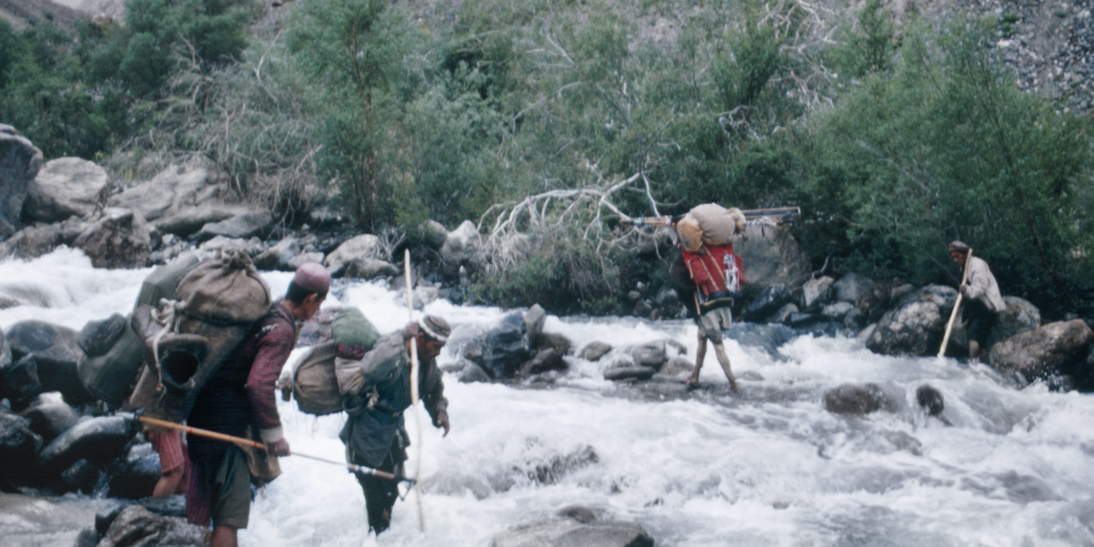 Four people walk through a rushing river. They are carrying heavy loads and using sticks for balance.