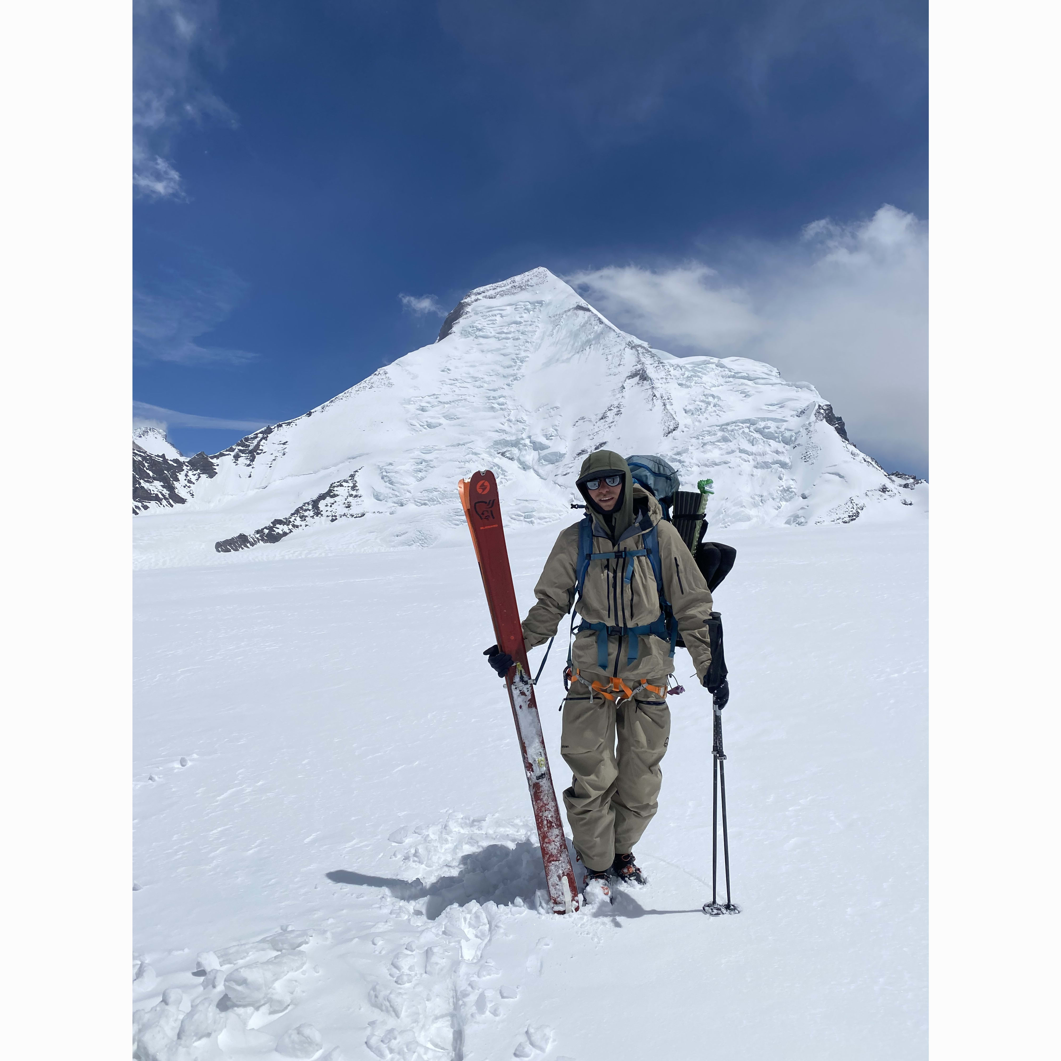 A skier on a snowy mountain wearing Norrøna clothing. They are equipped with skis and poles against a blue sky backdrop.