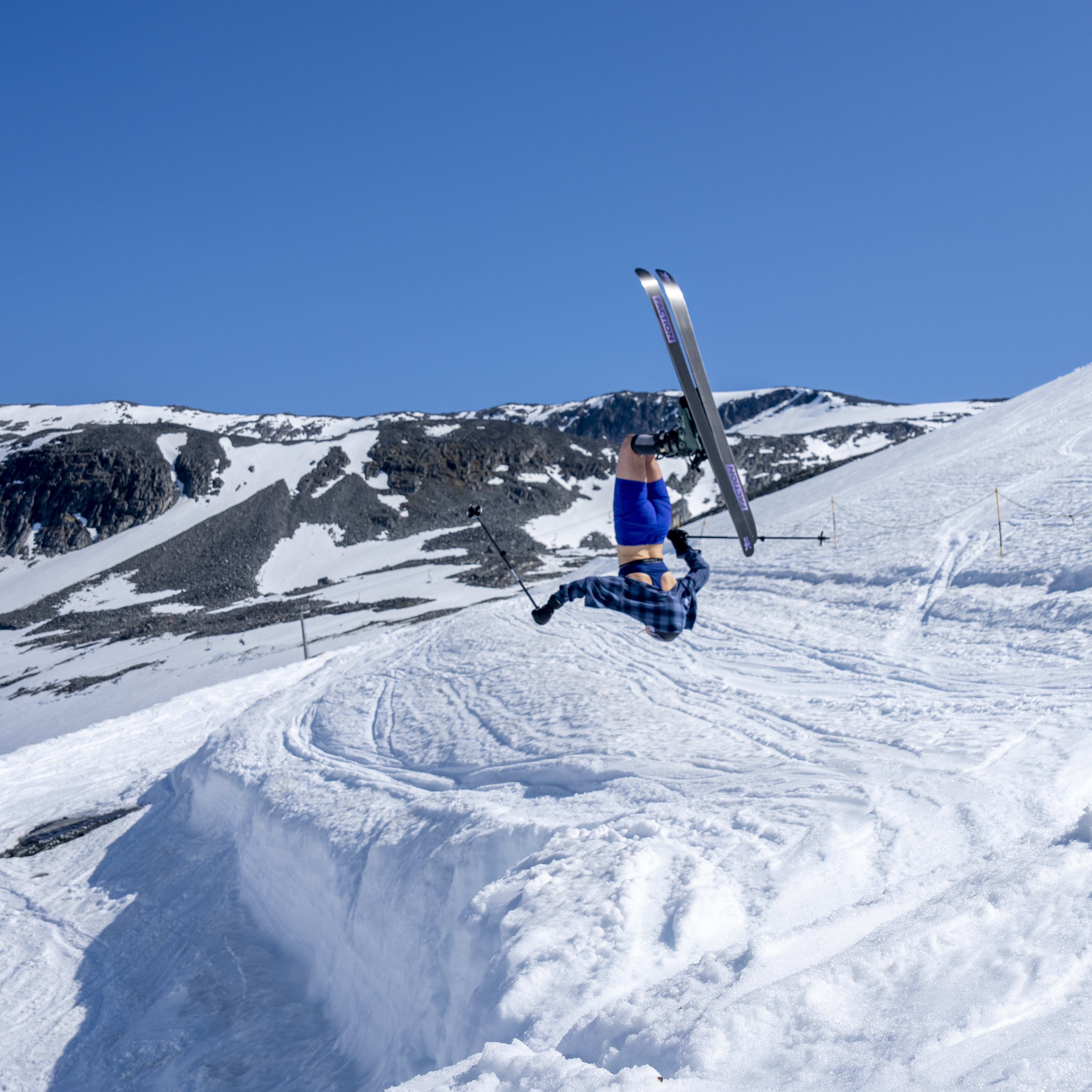 A skier wearing a blue Norrøna shirt and blue Norrøna shorts is upside down in the air while skiing on a snowy slope. The skis are from Norrøna.
