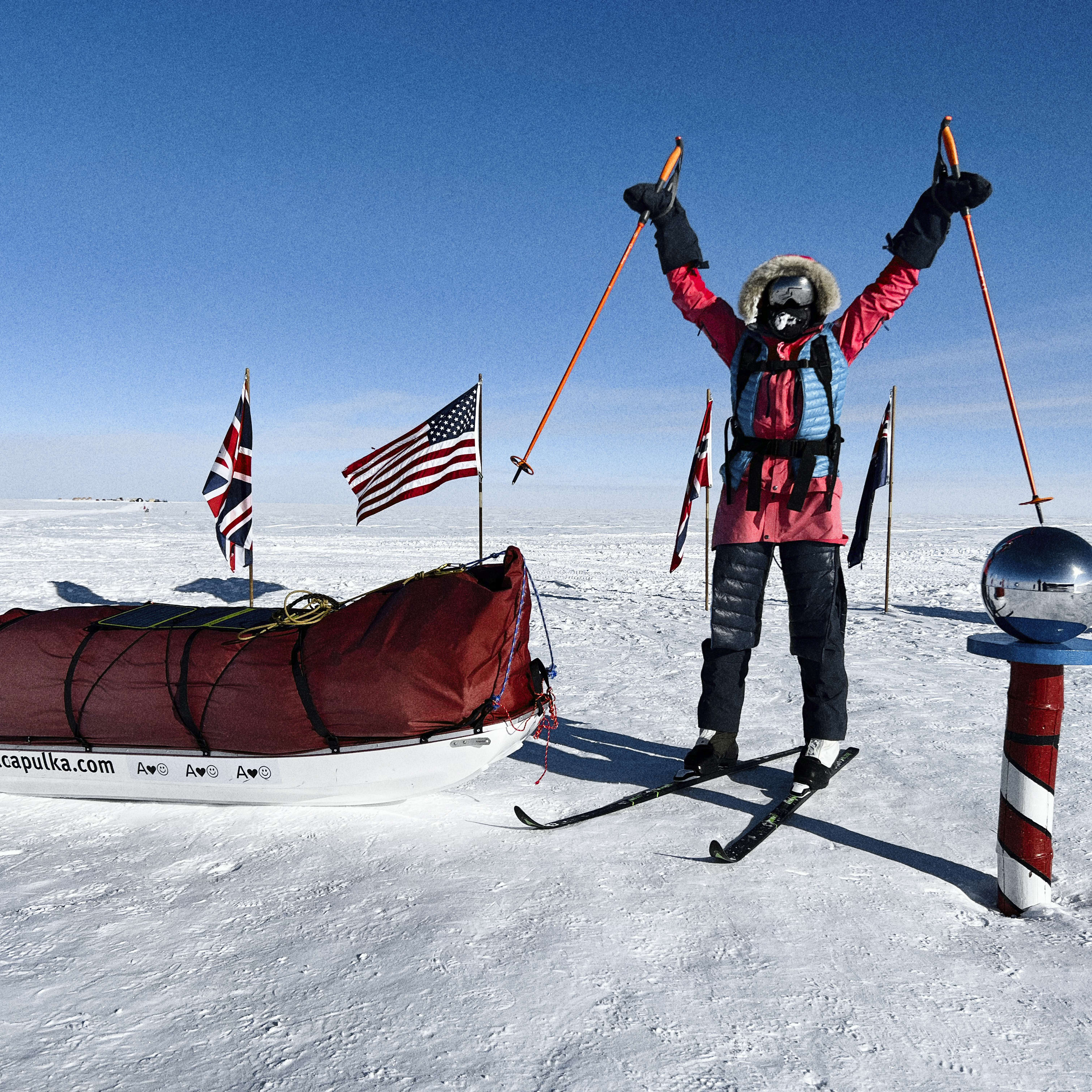 A person is standing in the snow with their arms raised holding ski poles, a red and white striped pole is beside them and a sled is behind them. There are flags in the distance.